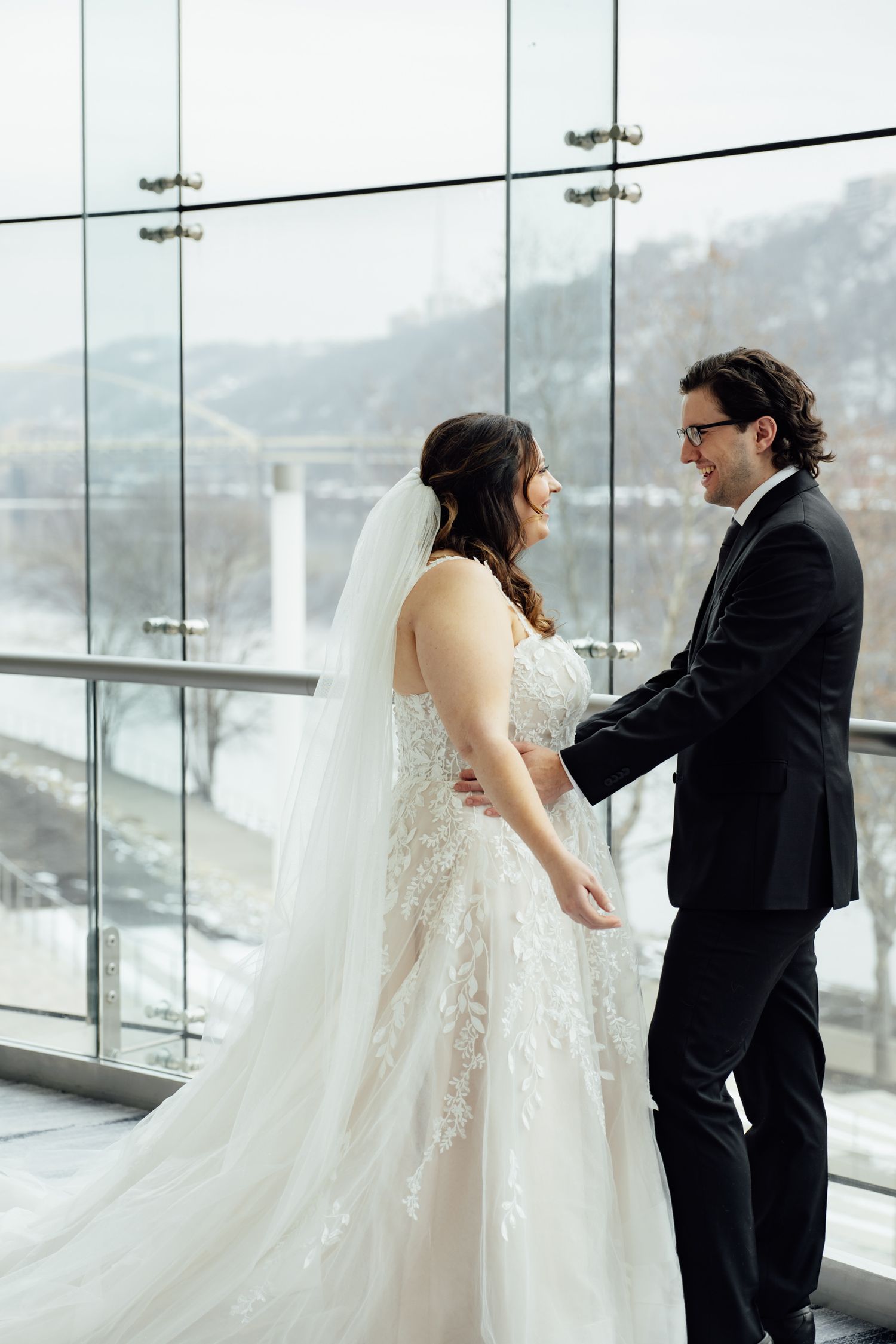 Bride and groom share a romantic moment by floor-to-ceiling windows with a scenic winter river view backdrop.