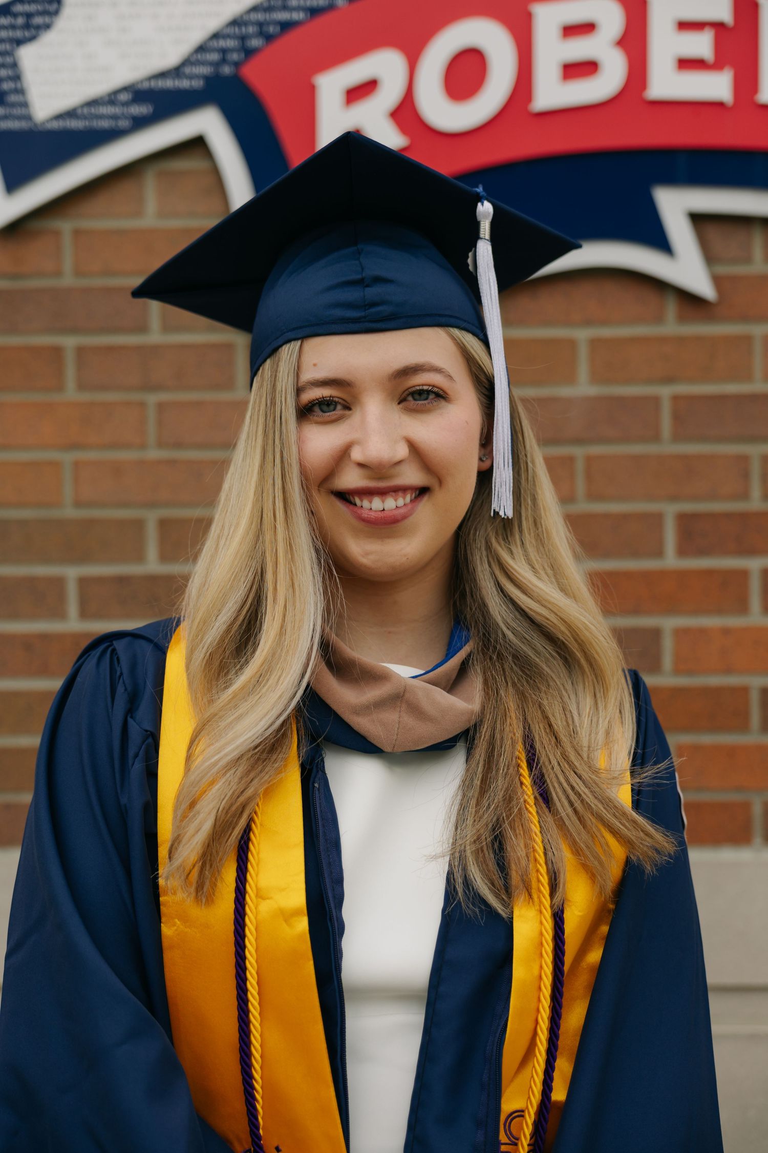 Smiling female graduate in navy cap and gown with gold stole poses in front of brick wall with school letters.