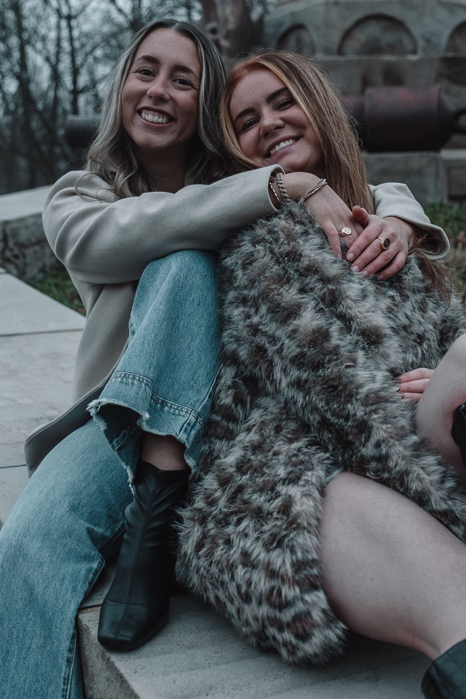 Two smiling young women sitting outdoors, one in jeans and boots, the other in a leopard print fur coat.
