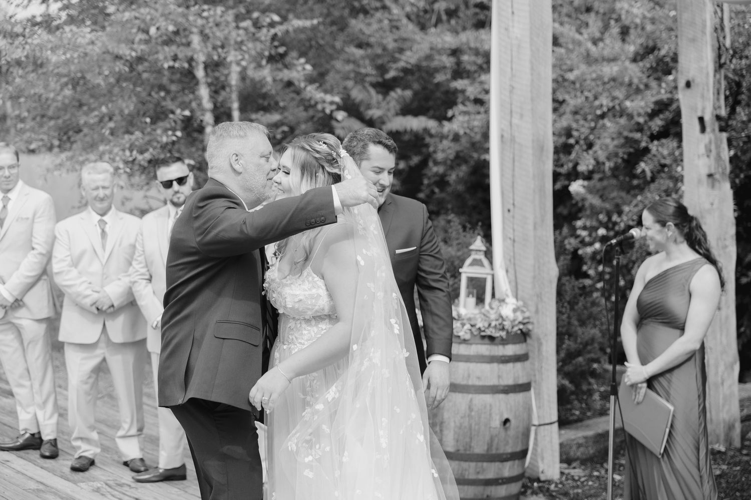Black and white photo of a groom kissing his bride at an outdoor rustic wedding ceremony.