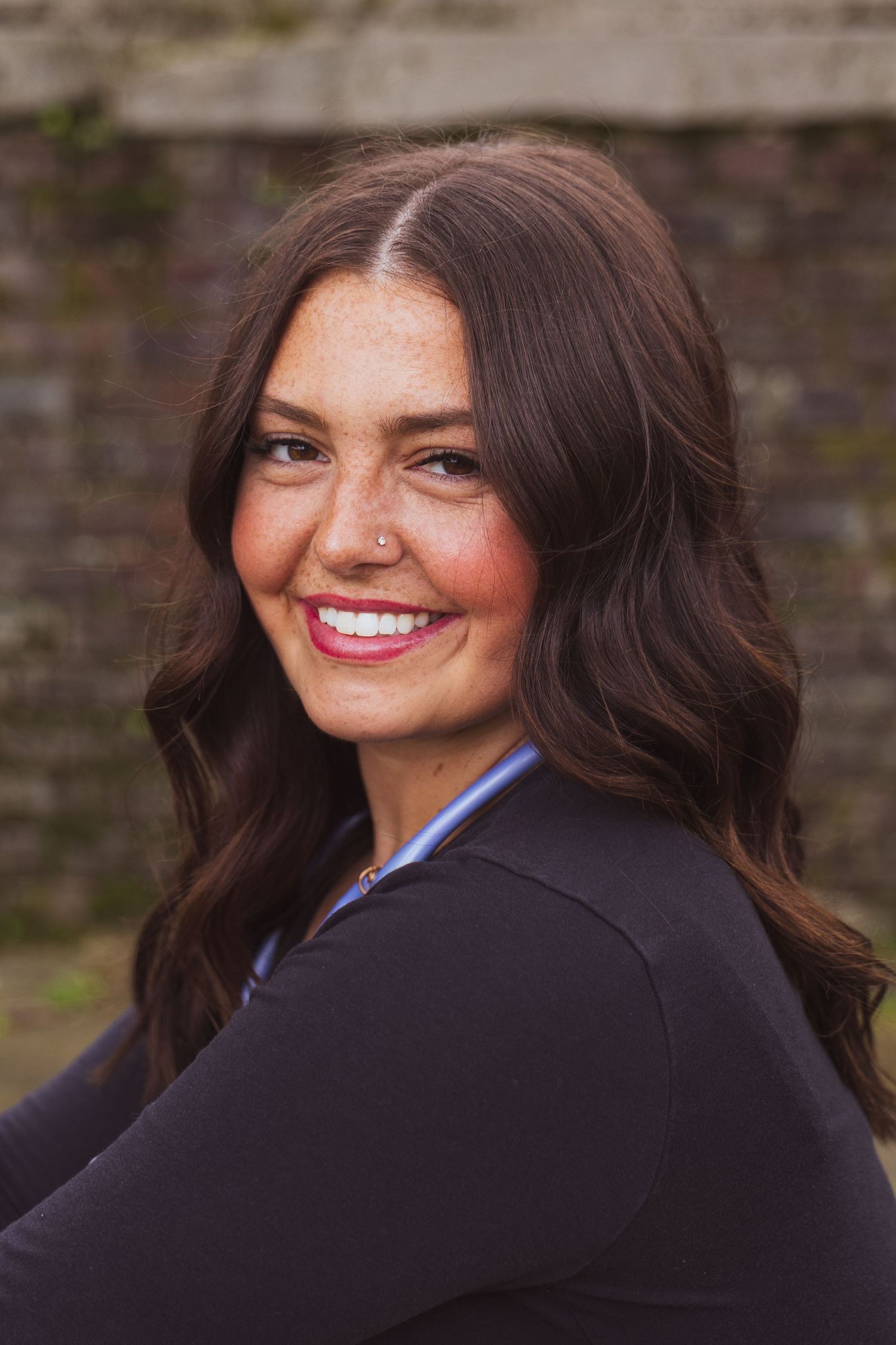 A woman with long dark wavy hair and bright smile wearing a black blazer poses for a professional headshot outdoors.