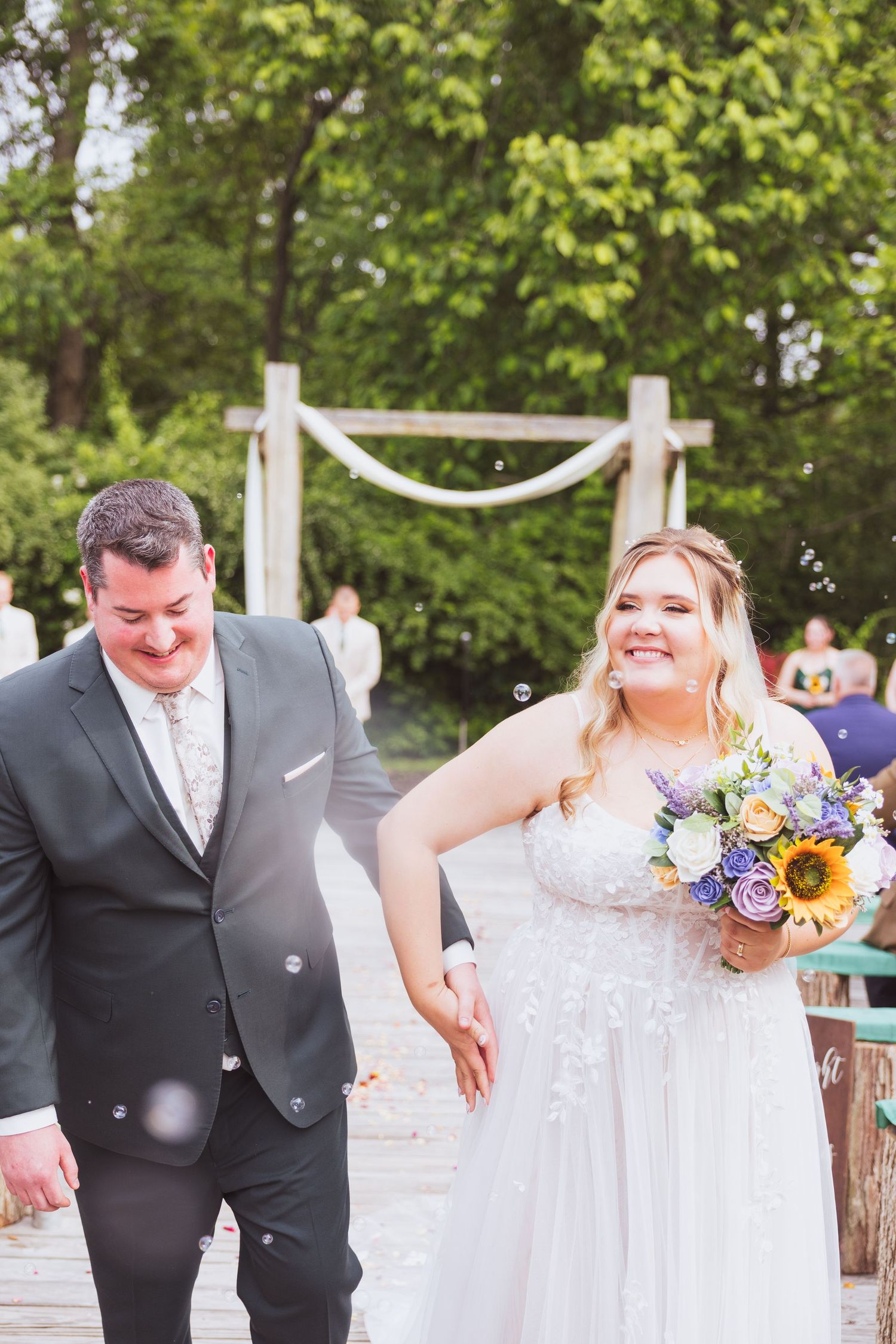 Joyful wedding couple walking down aisle after ceremony under white draped arch with sunflowers in bouquet.