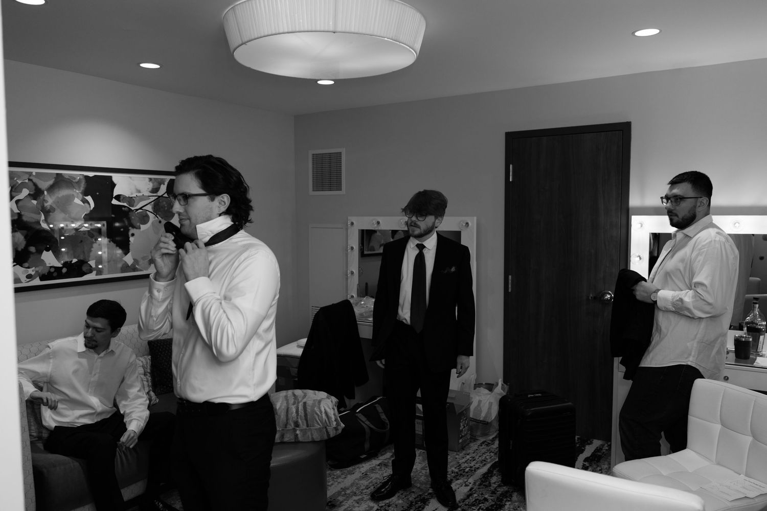 Black and white photo of groomsmen getting ready in a hotel room, adjusting ties and suits before a wedding.