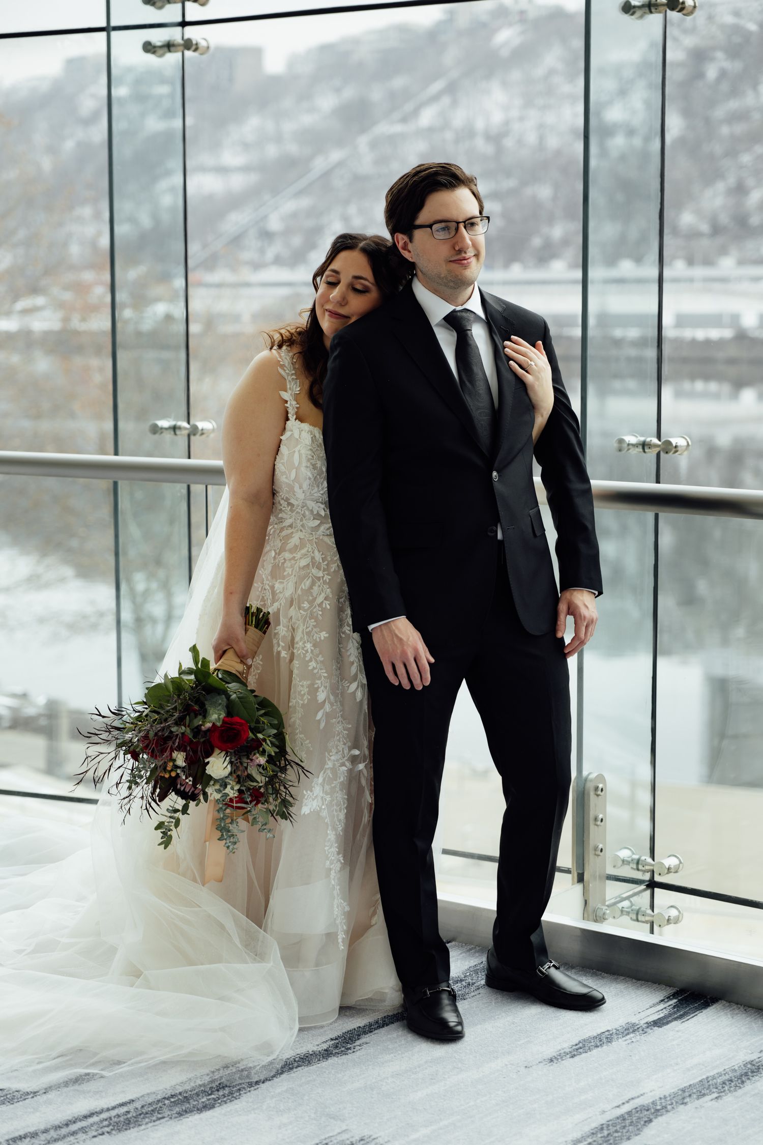 Bride in lace gown leans on groom in black suit, holding red bouquet, with snowy mountain backdrop.
