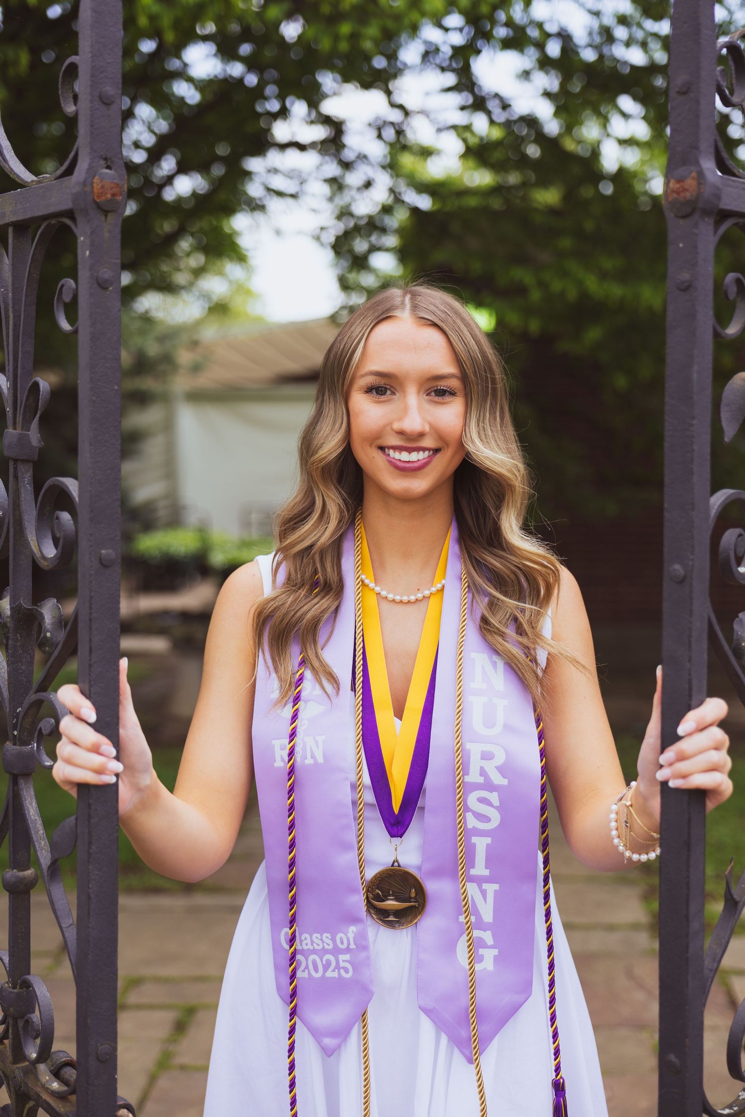 Graduate wearing white dress with purple graduation stole and gold medals poses at decorative metal gate outdoors.