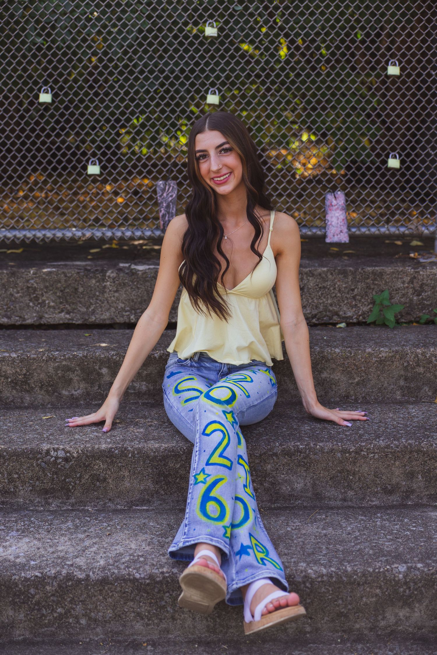 Young woman smiling in yellow top and colorful number-print flare jeans, sitting on stone steps outdoors.