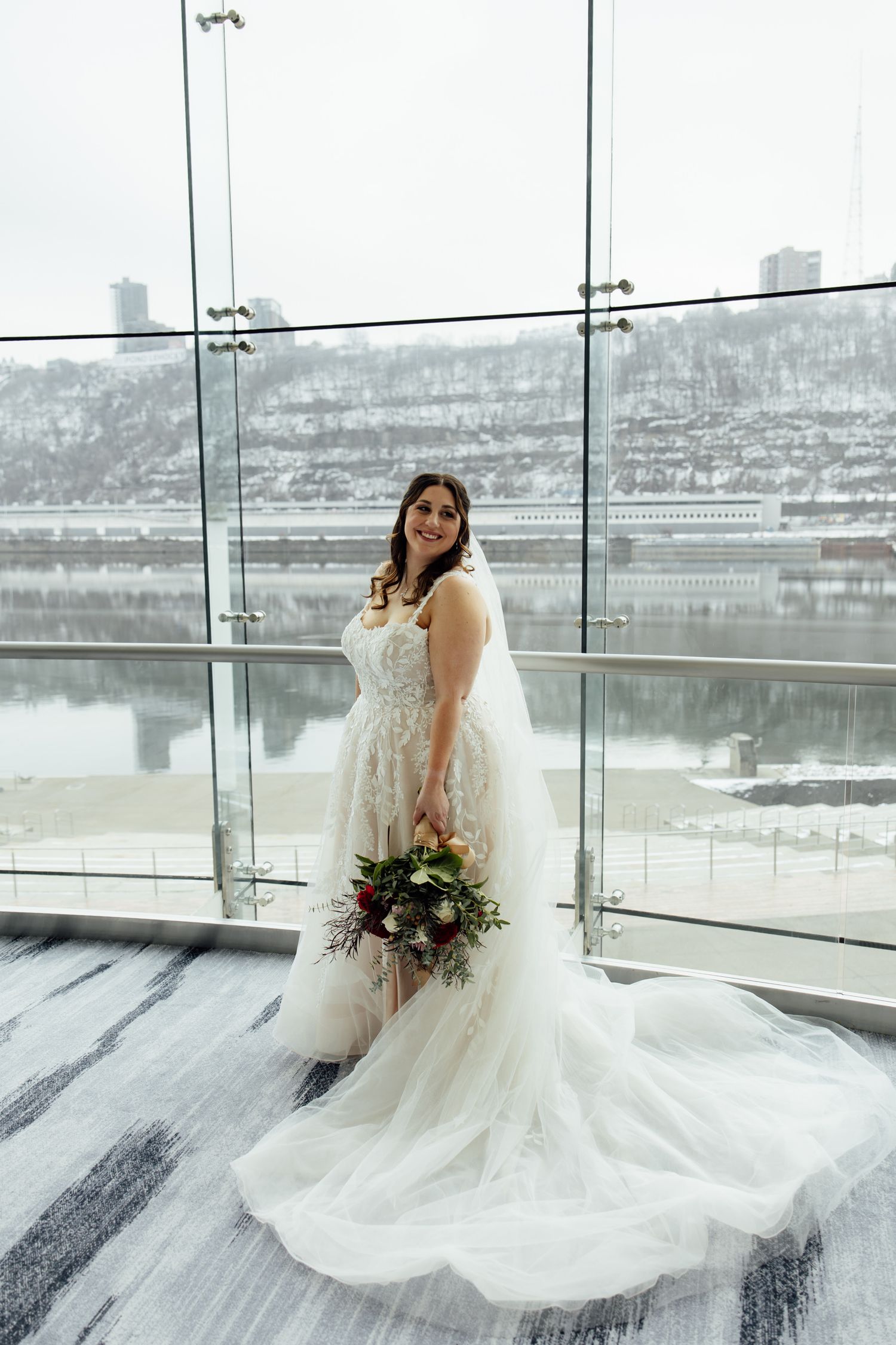 Bride in lace wedding gown holding bouquet poses by floor-to-ceiling windows with snowy river view.