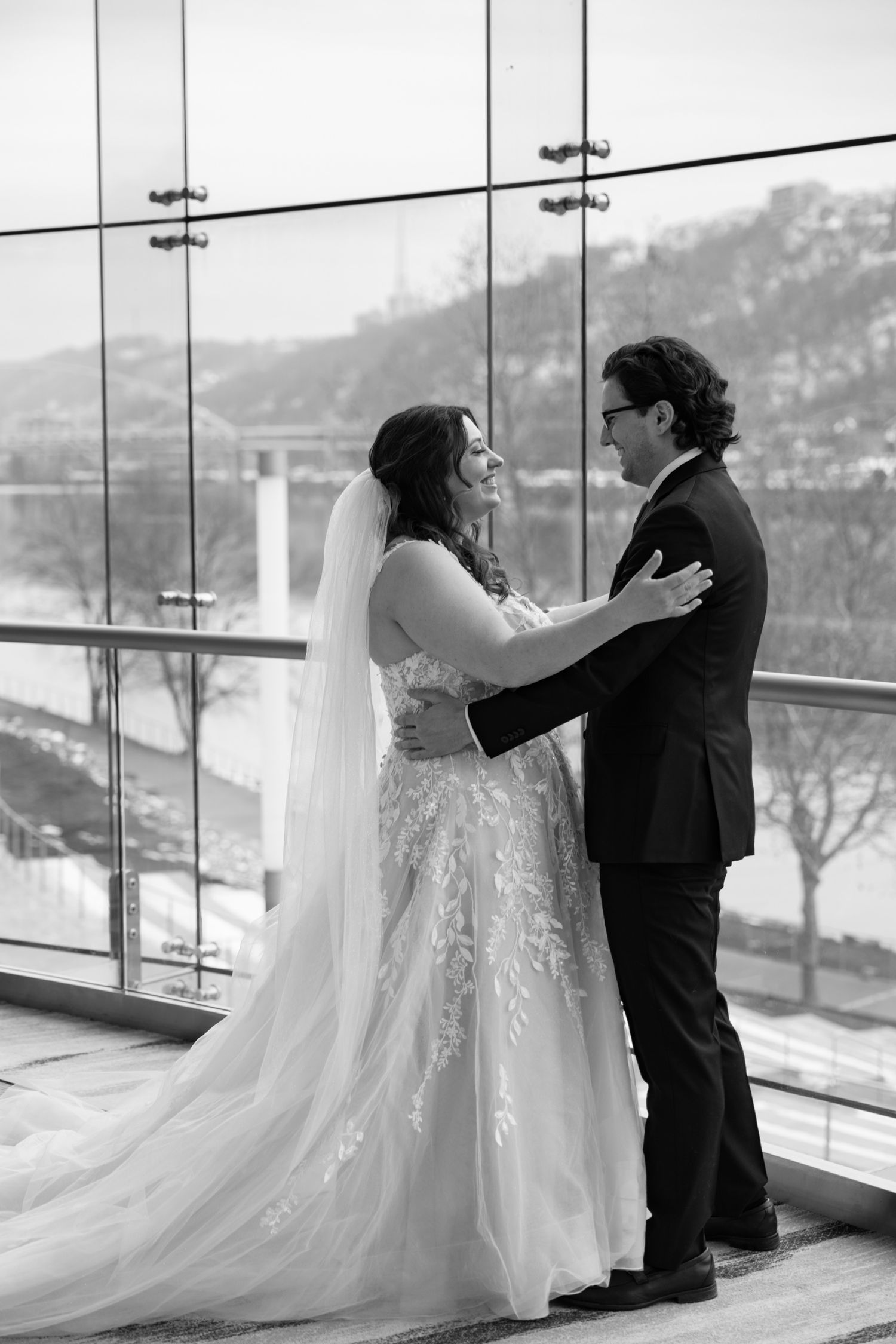 Bride and groom share tender moment in black and white photo, embracing before large windows with scenic backdrop.