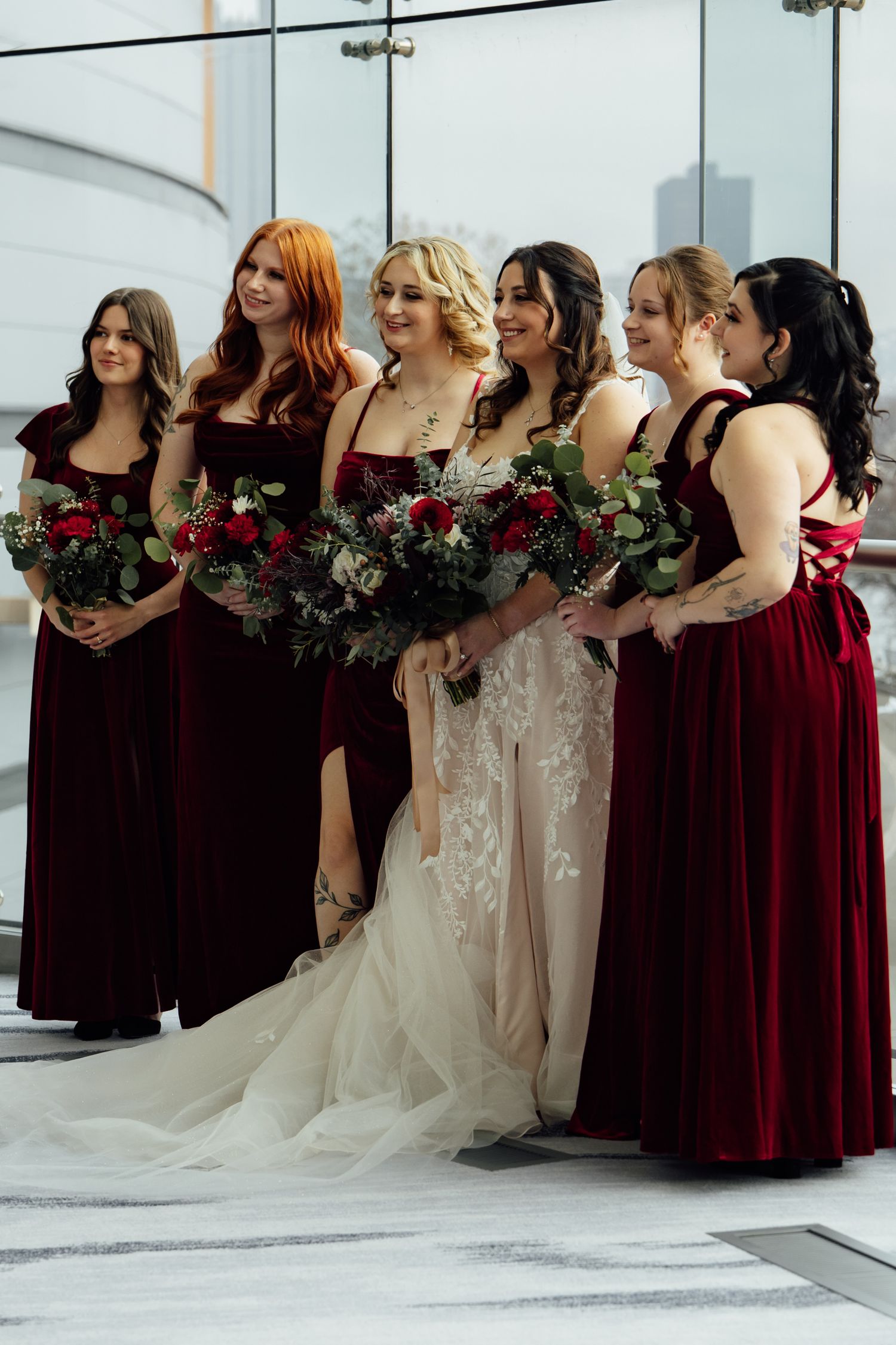 Bride in lace gown with five bridesmaids in burgundy velvet dresses holding red floral bouquets.