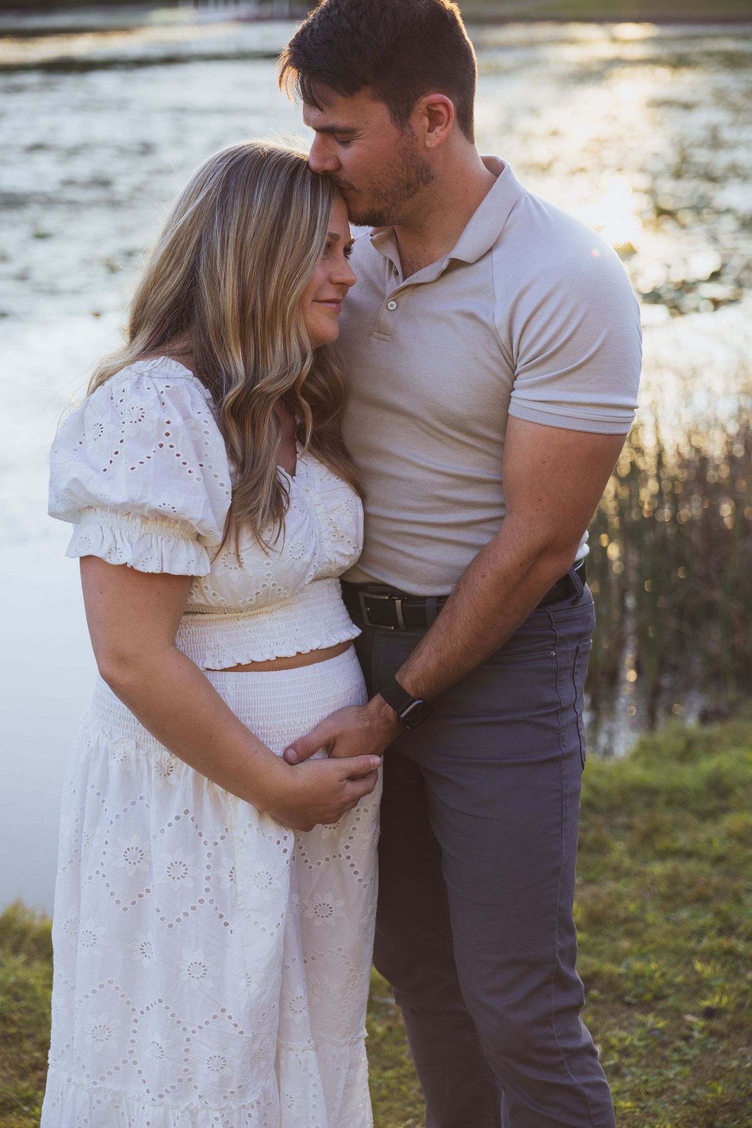 Couple sharing a tender moment outdoors by a lake, woman in white lace dress with visible baby bump, man in beige polo shirt.