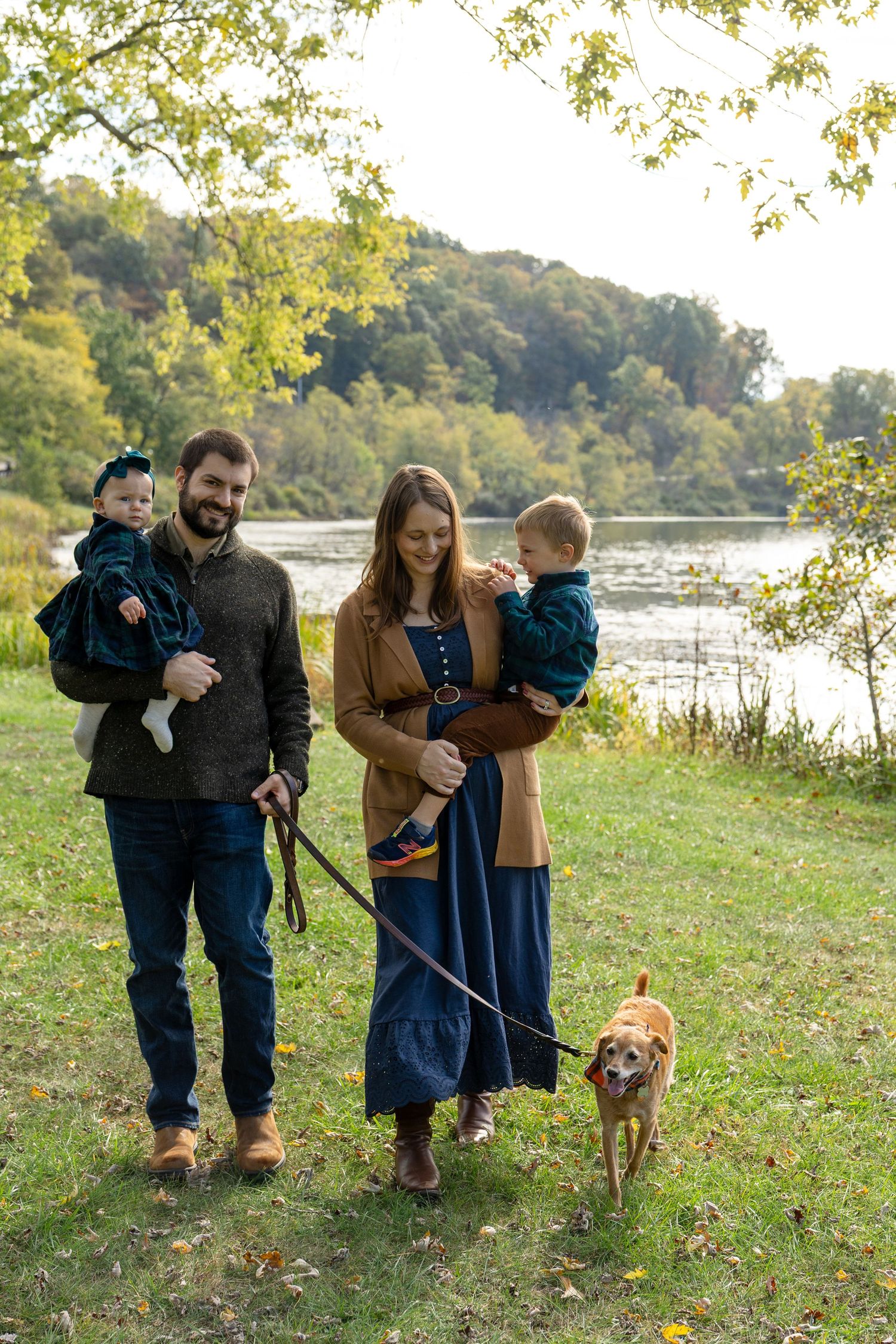 Family of two adults holding toddlers with a golden dog on leash, walking by a scenic river in autumn.