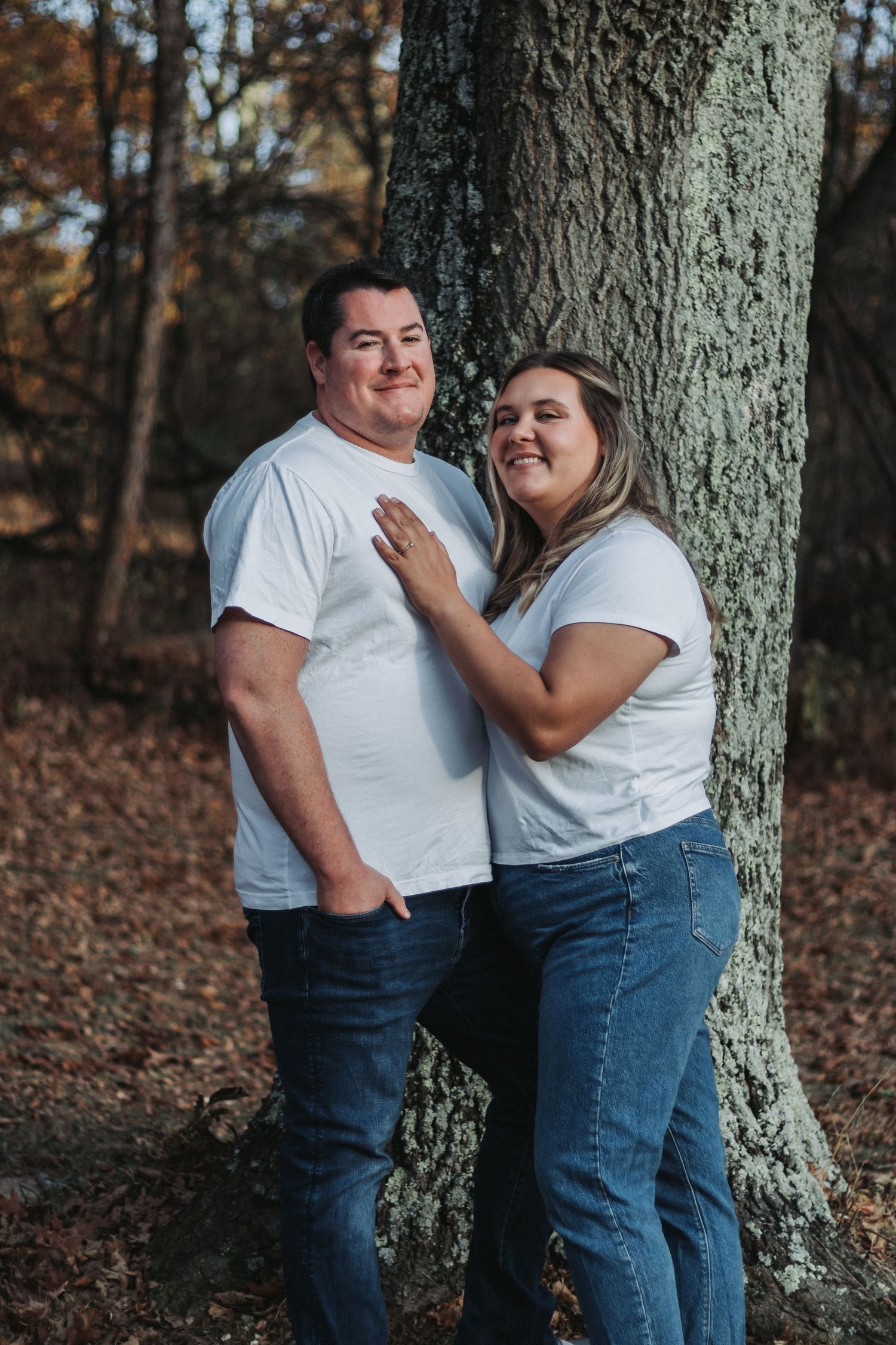 Couple in white shirts posing together against large tree trunk in autumn forest.