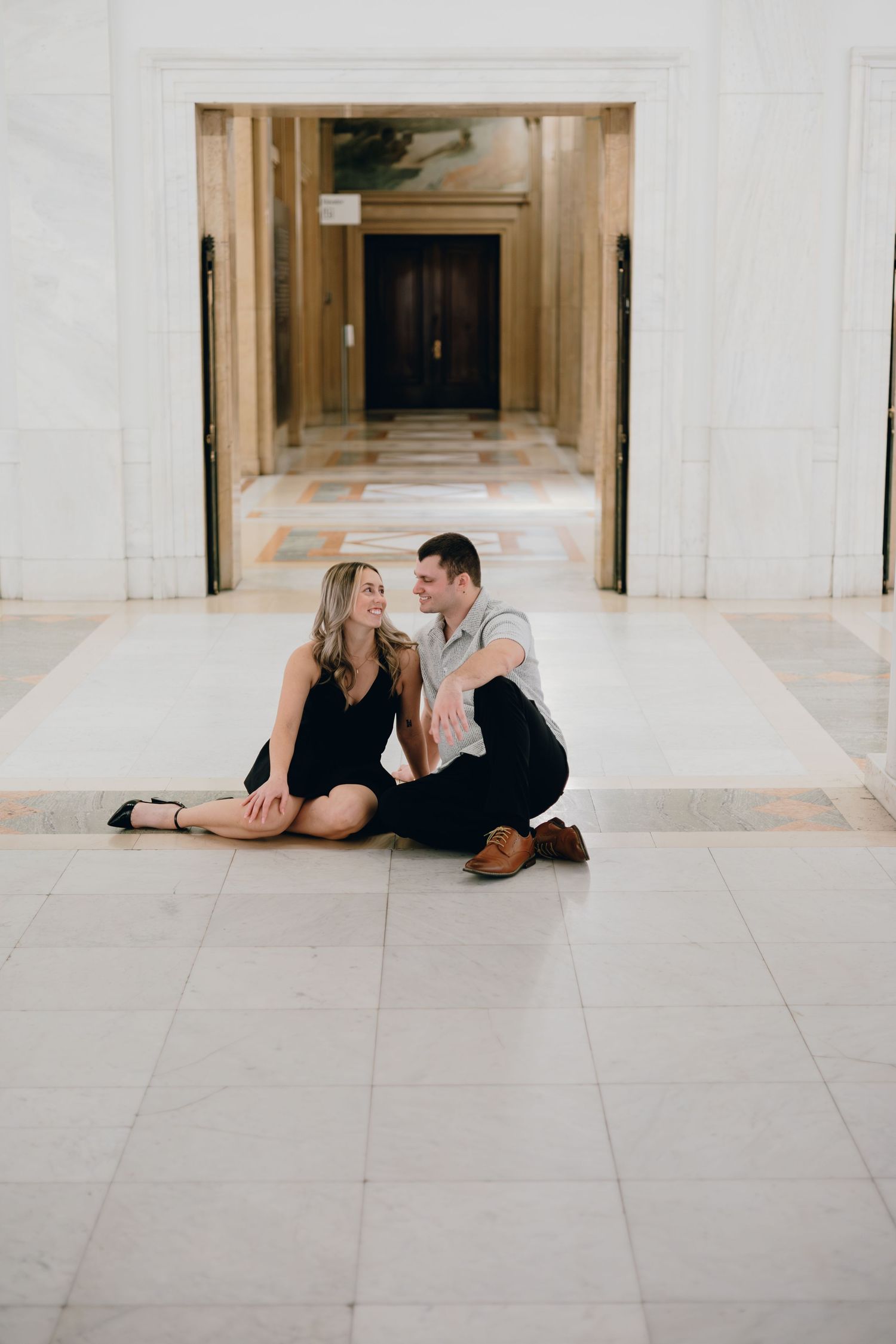 A couple sitting together on a marble floor in an elegant hallway with architectural columns.