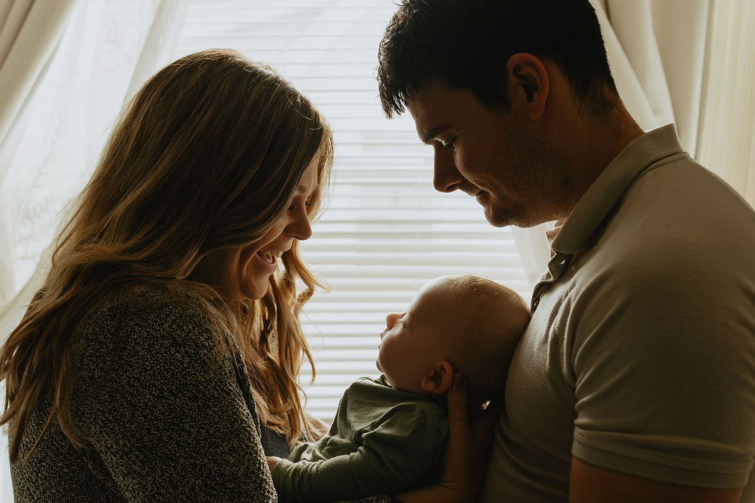 Young parents smiling lovingly at their newborn baby by a bright window in a warm, intimate family portrait.