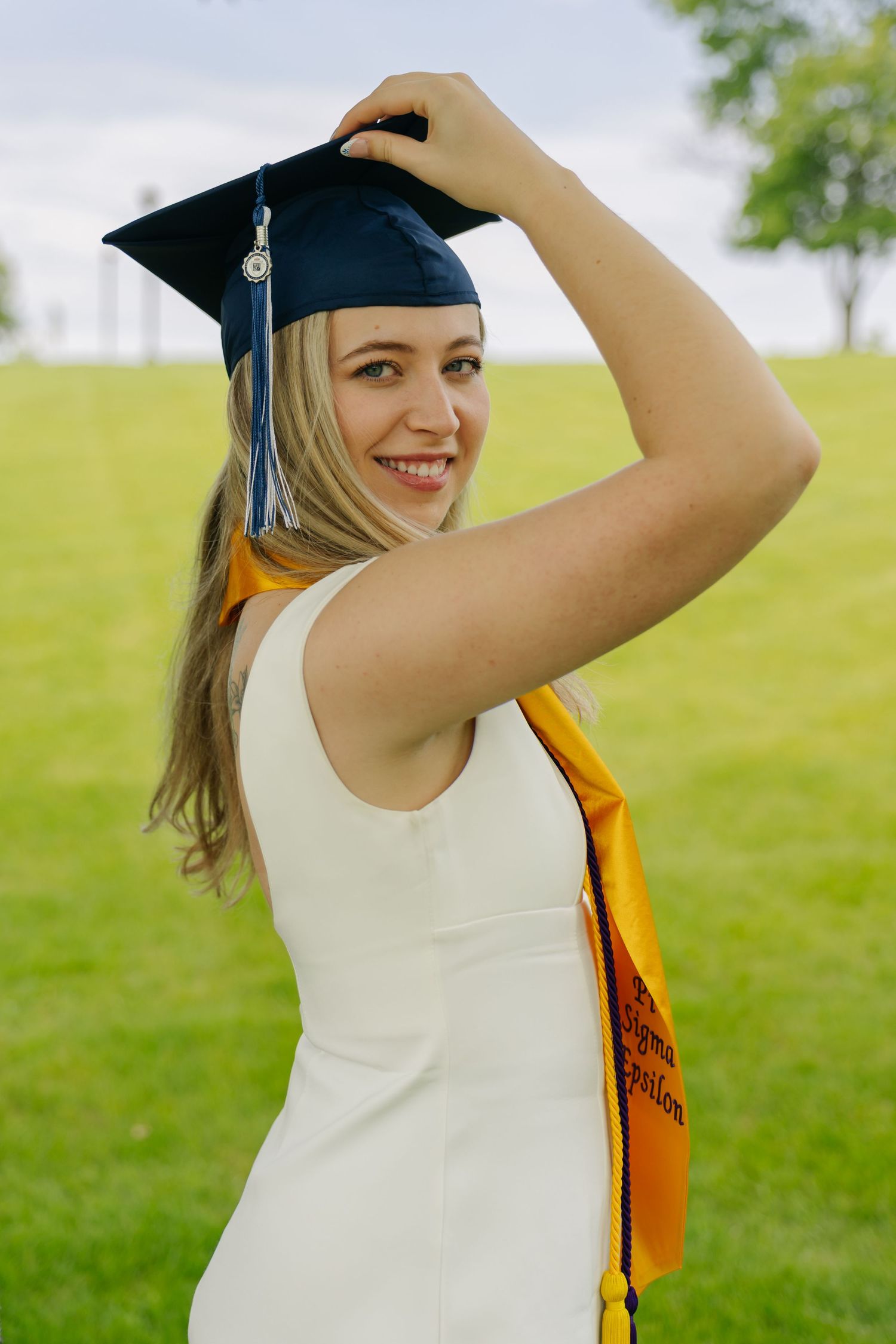 Smiling blonde woman in white dress wearing graduation cap and gold honor stole outdoors on green lawn.