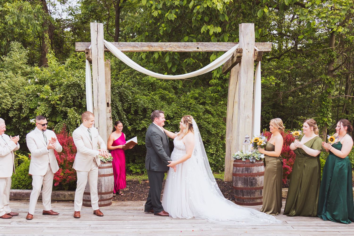 Bride and groom exchange vows under rustic wooden arch with draped fabric at outdoor wedding ceremony.