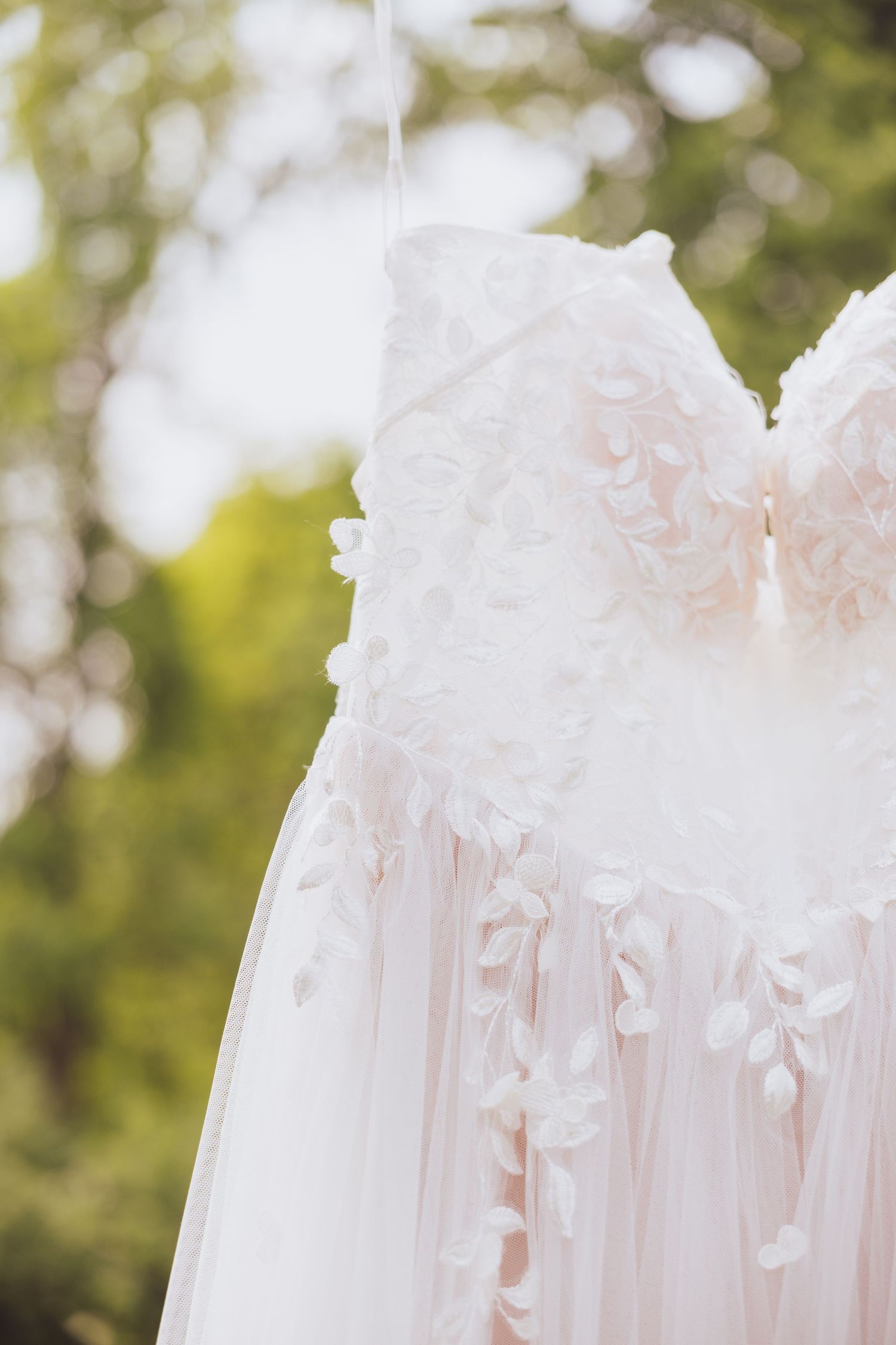 Close-up detail of delicate white lace and tulle wedding dress bodice hanging outdoors with soft bokeh background.