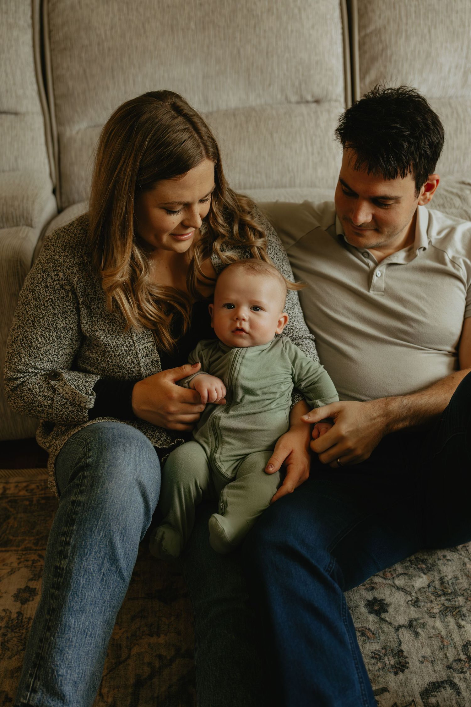 Young family with mother, father, and baby in green onesie sitting together on couch at home.