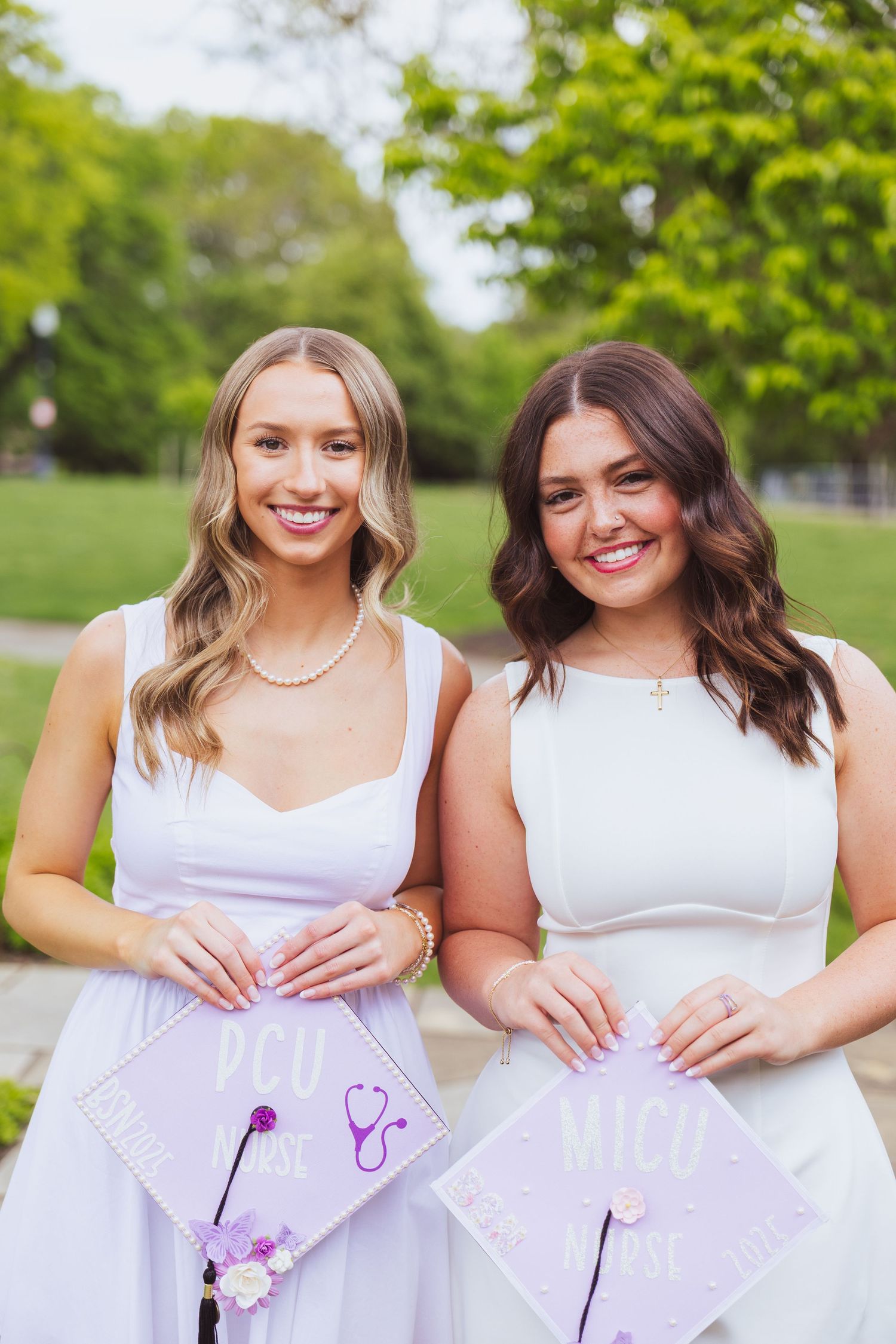 Two graduates in white dresses pose together outdoors while holding decorated graduation caps.