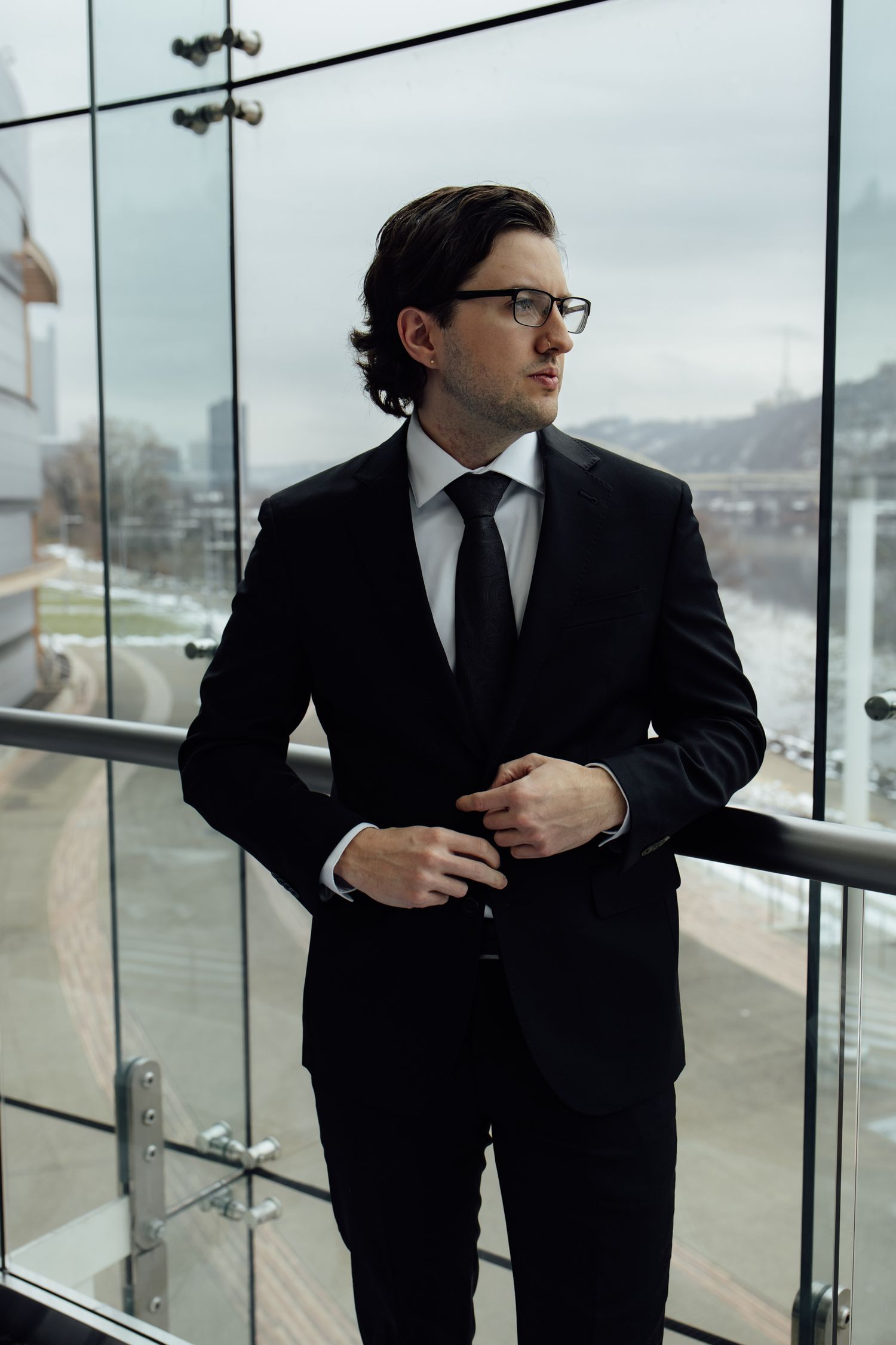 Professional businessman in black suit and tie adjusting jacket on modern glass balcony.