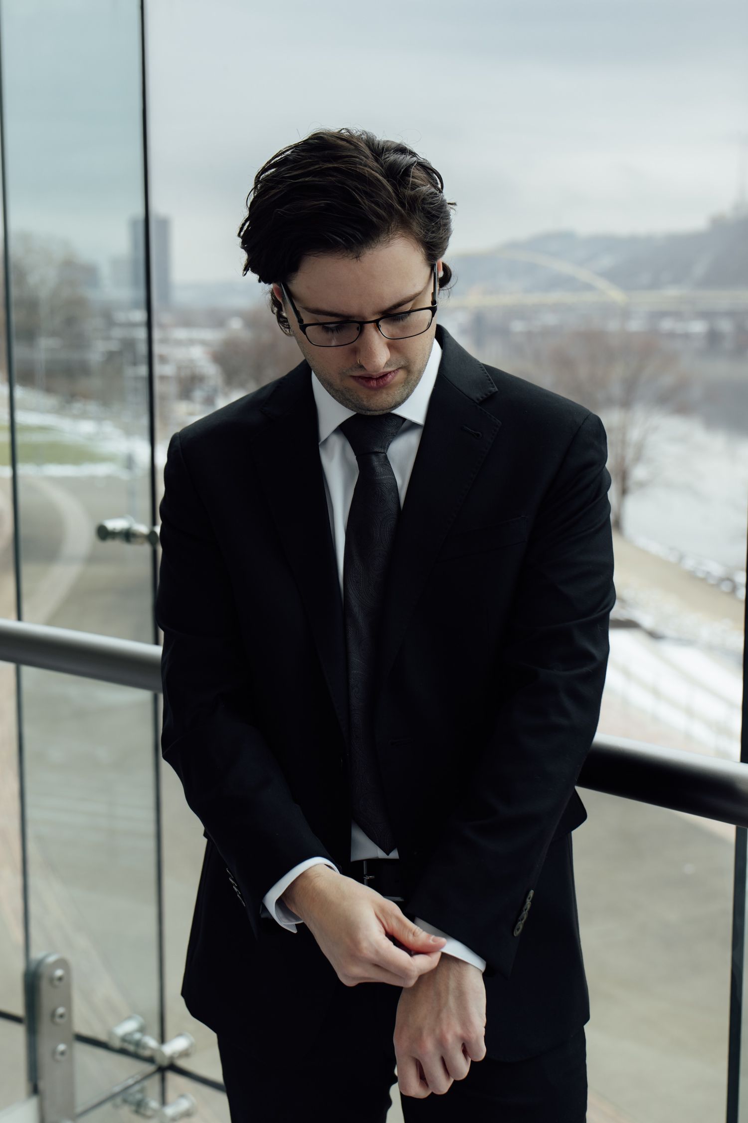 A young businessman in a black suit and tie adjusts his cufflinks on a glass balcony overlooking a city river.