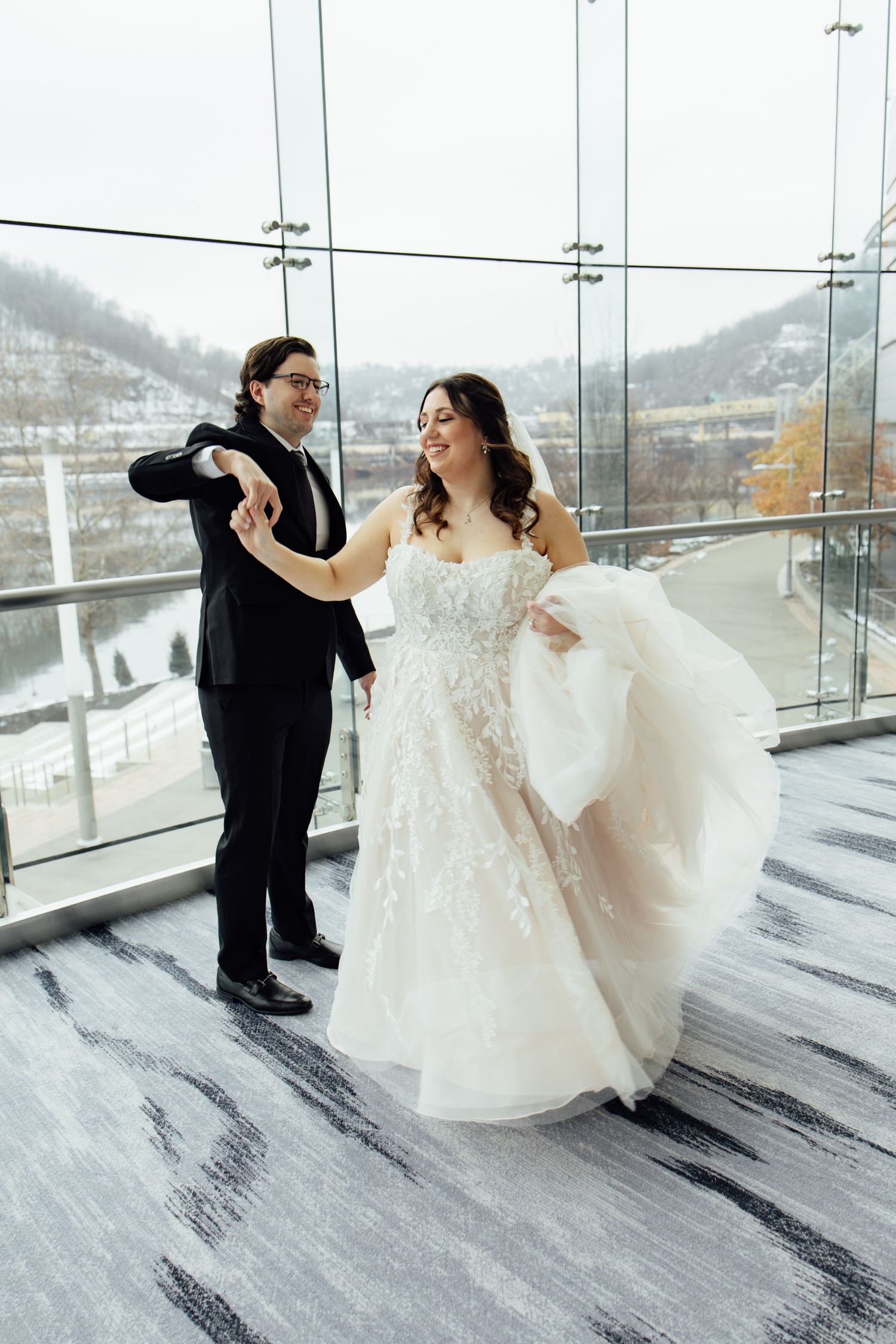 Bride and groom dancing joyfully in elegant wedding attire near large glass windows with scenic winter view.
