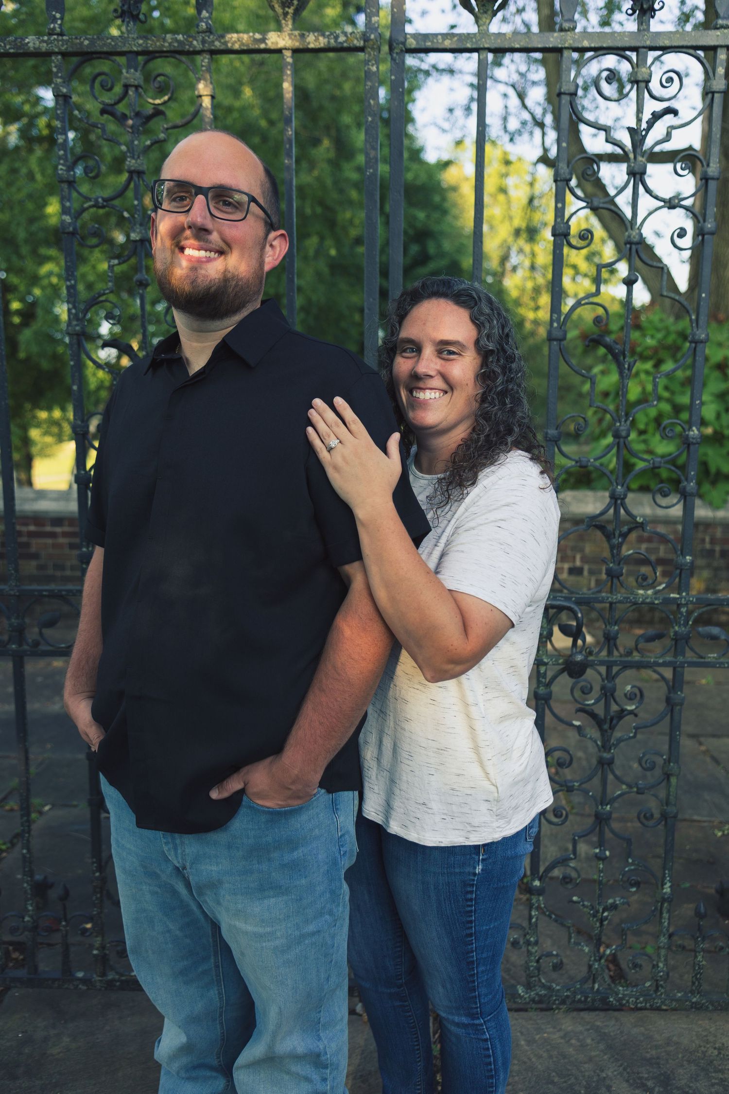 Couple posing in front of ornate iron gate, woman showing engagement ring while embracing man in black shirt and jeans.