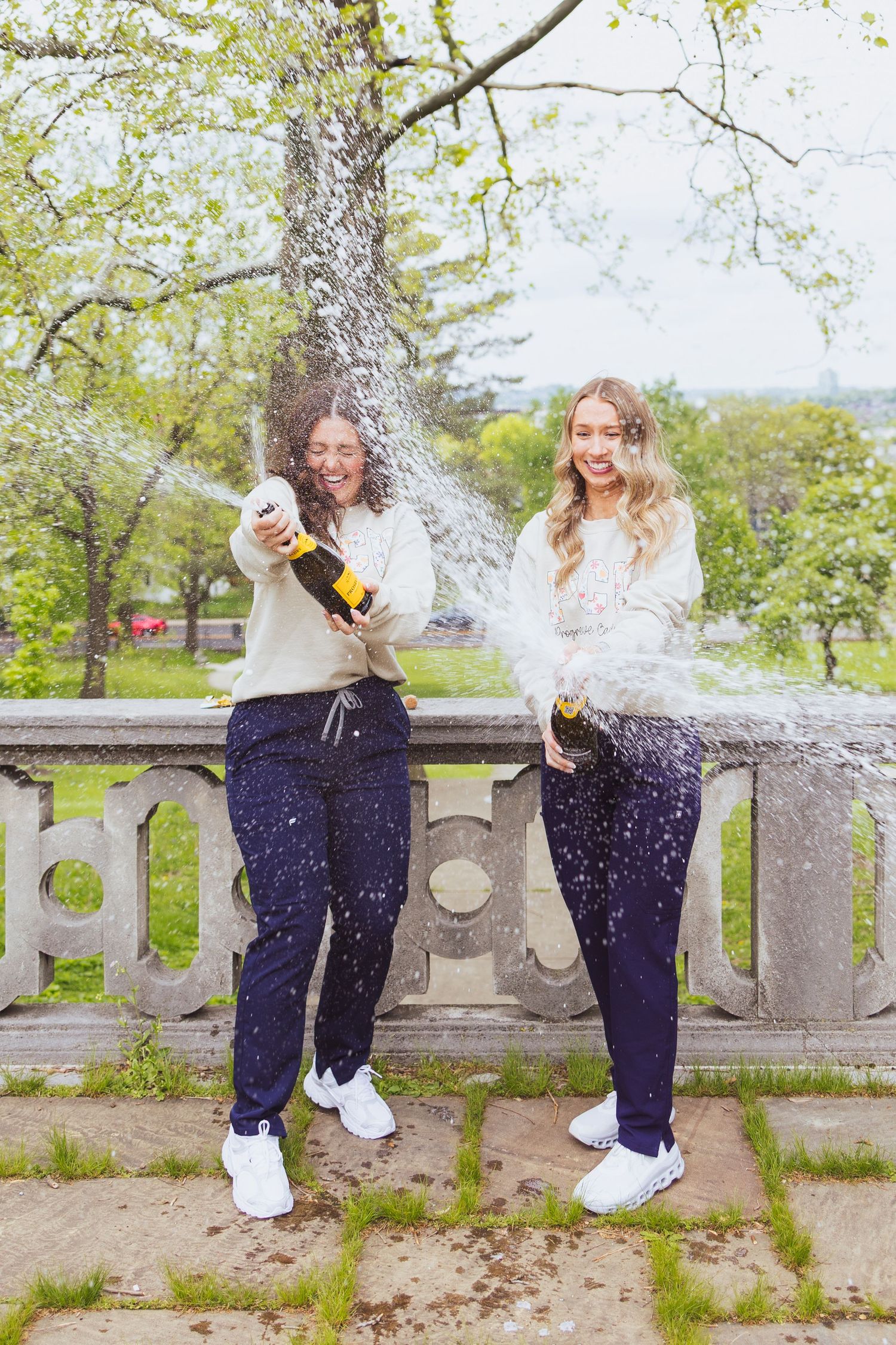 Two friends celebrating with champagne spraying on a stone balcony surrounded by green trees on a spring day.