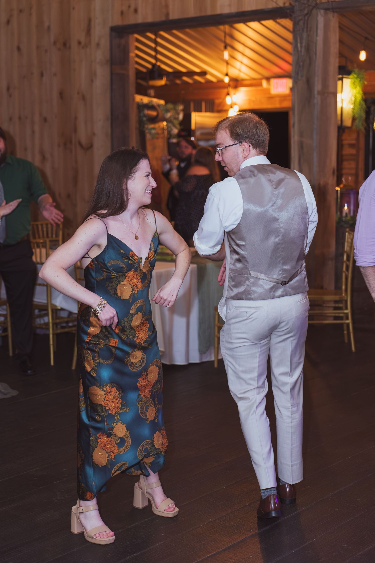 Couple dancing together at a rustic barn wedding reception, woman in floral dress, man in vest and slacks.