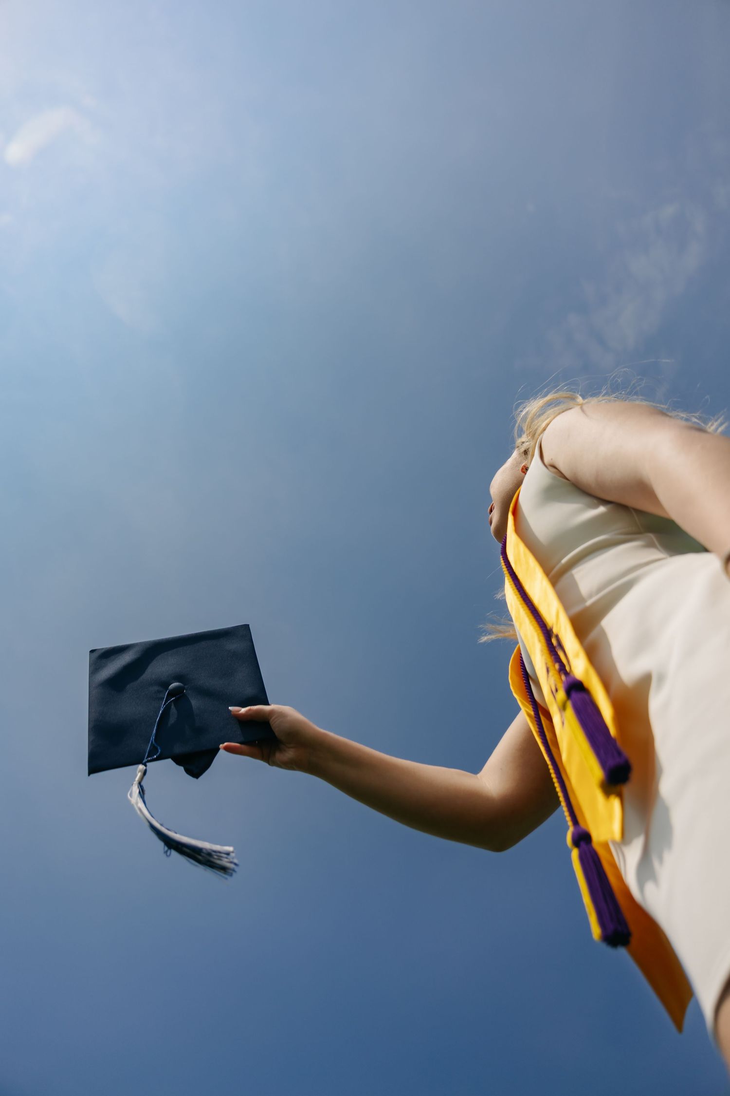 Graduate holding mortarboard cap against blue sky, wearing yellow and purple academic regalia at outdoor ceremony.