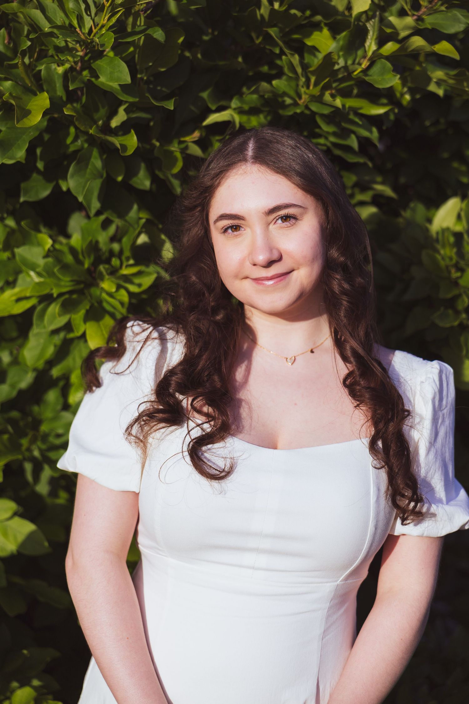 A portrait outdoors featuring long dark curly hair, white dress with puff sleeves against a leafy green background.