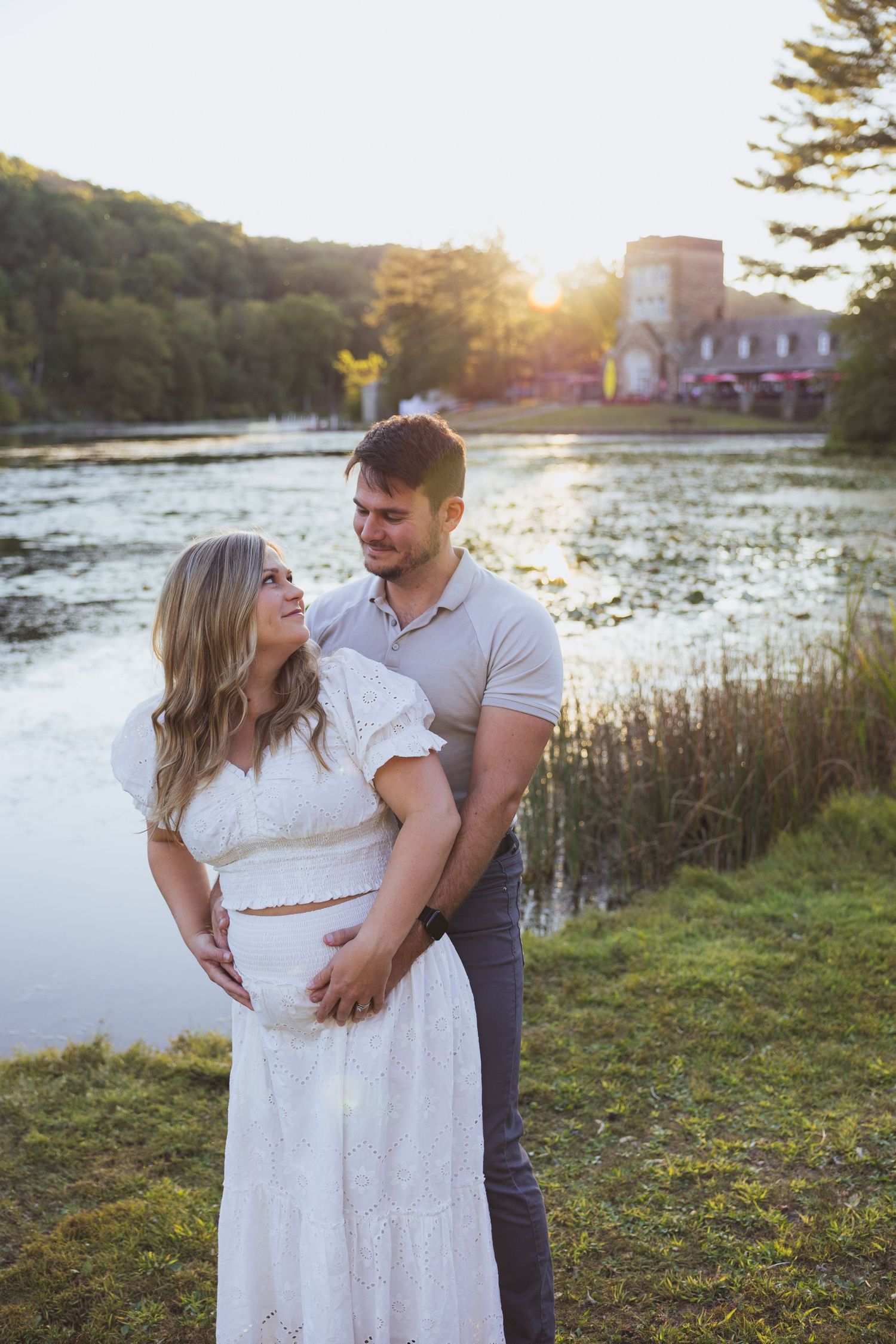 Couple embracing by a sunlit lake at golden hour, woman in white dress with man in gray shirt, lush greenery background.