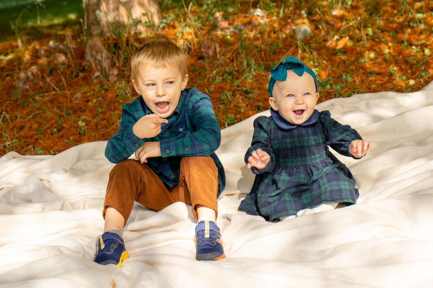 Two happy young children sitting on a white blanket outdoors in autumn, boy in plaid shirt and girl in teal dress.