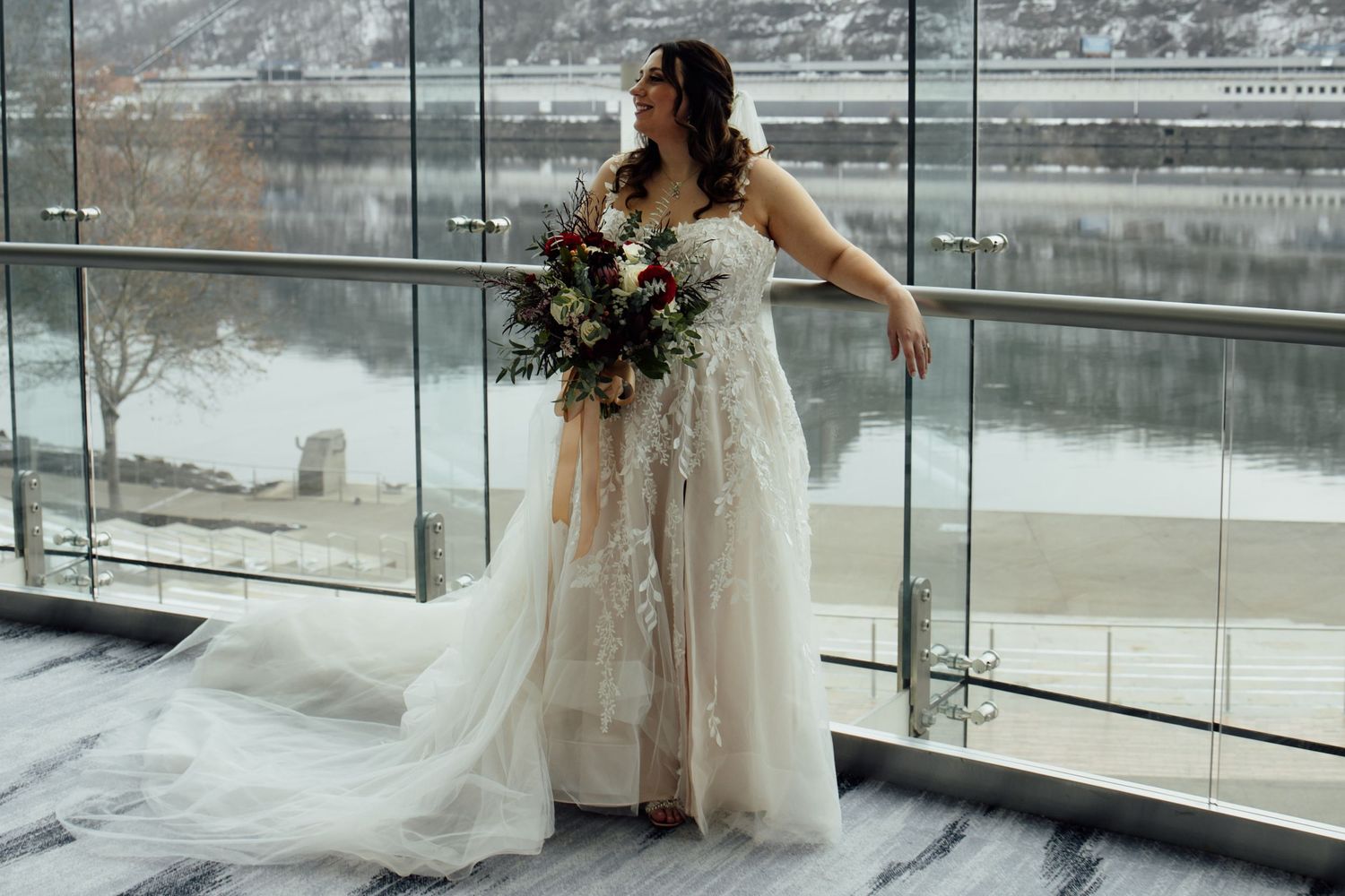 Bride in lace wedding gown holding floral bouquet poses by glass railing with snowy river view.