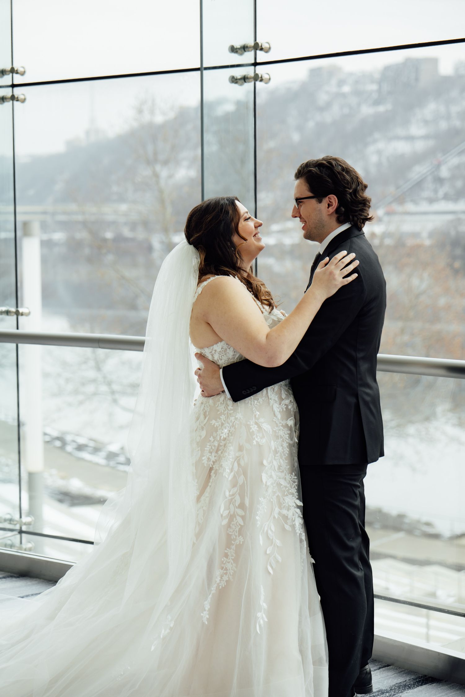 Bride and groom embrace near large windows with snowy river view, bride in lace gown with veil, groom in black tuxedo.