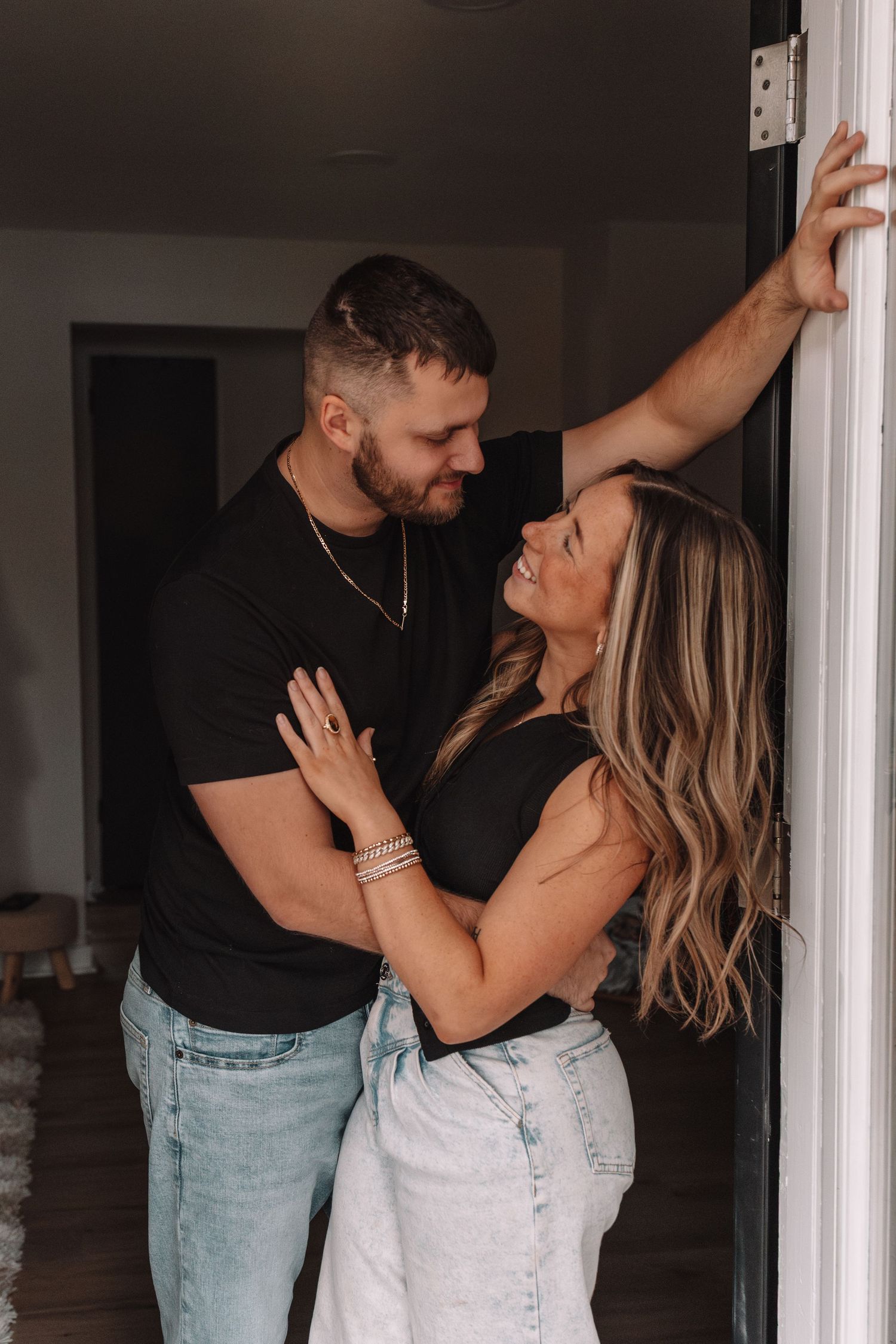 A couple in matching black tops and jeans share a romantic moment in a doorway, smiling at each other.