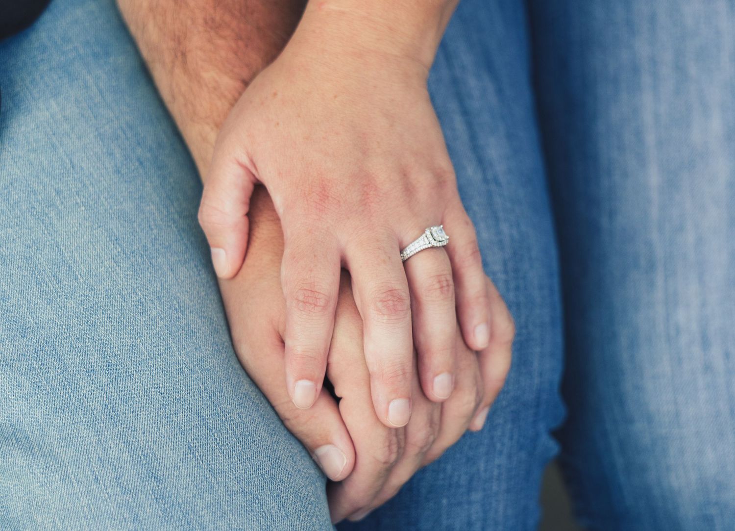 Close-up of a couple holding hands, woman wearing a diamond engagement ring, both dressed in blue jeans.