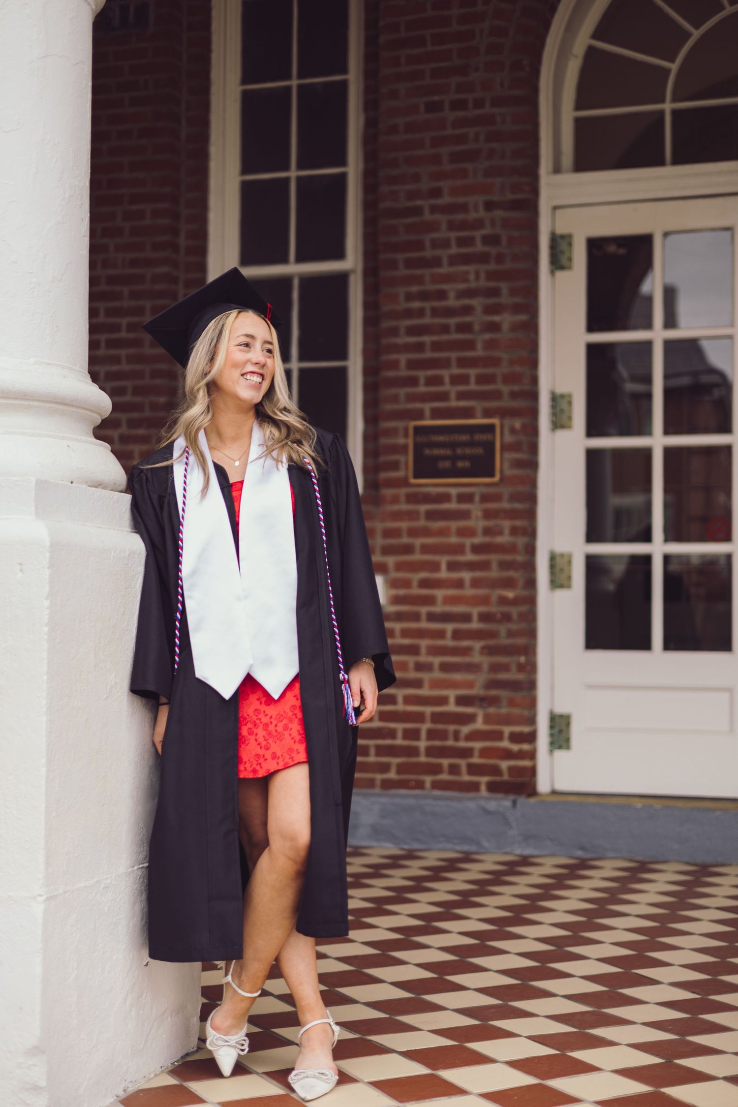 Graduate in academic regalia poses by brick building entrance with checkered flooring.