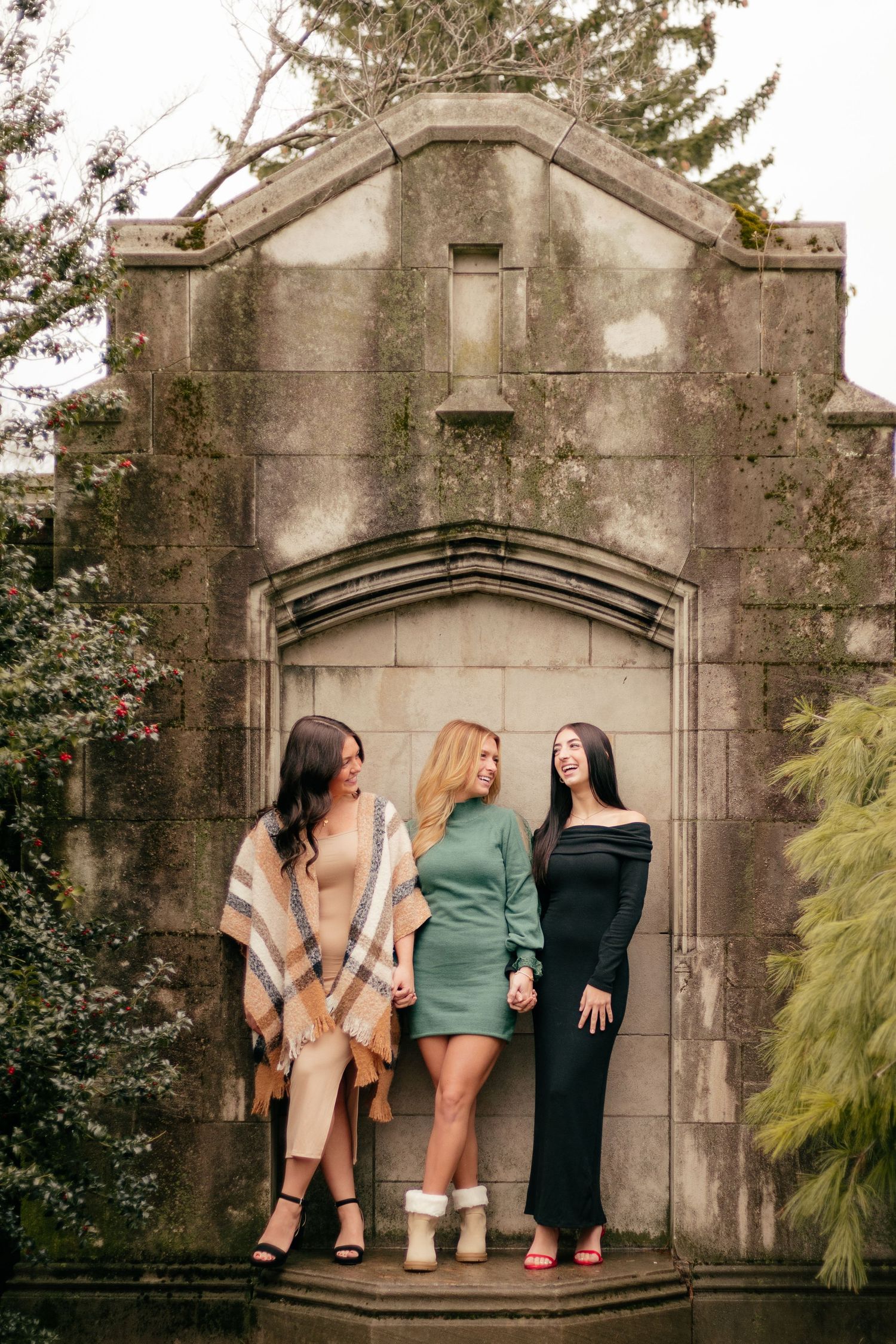 Three women in stylish outfits standing on stone steps against gothic architecture and greenery.