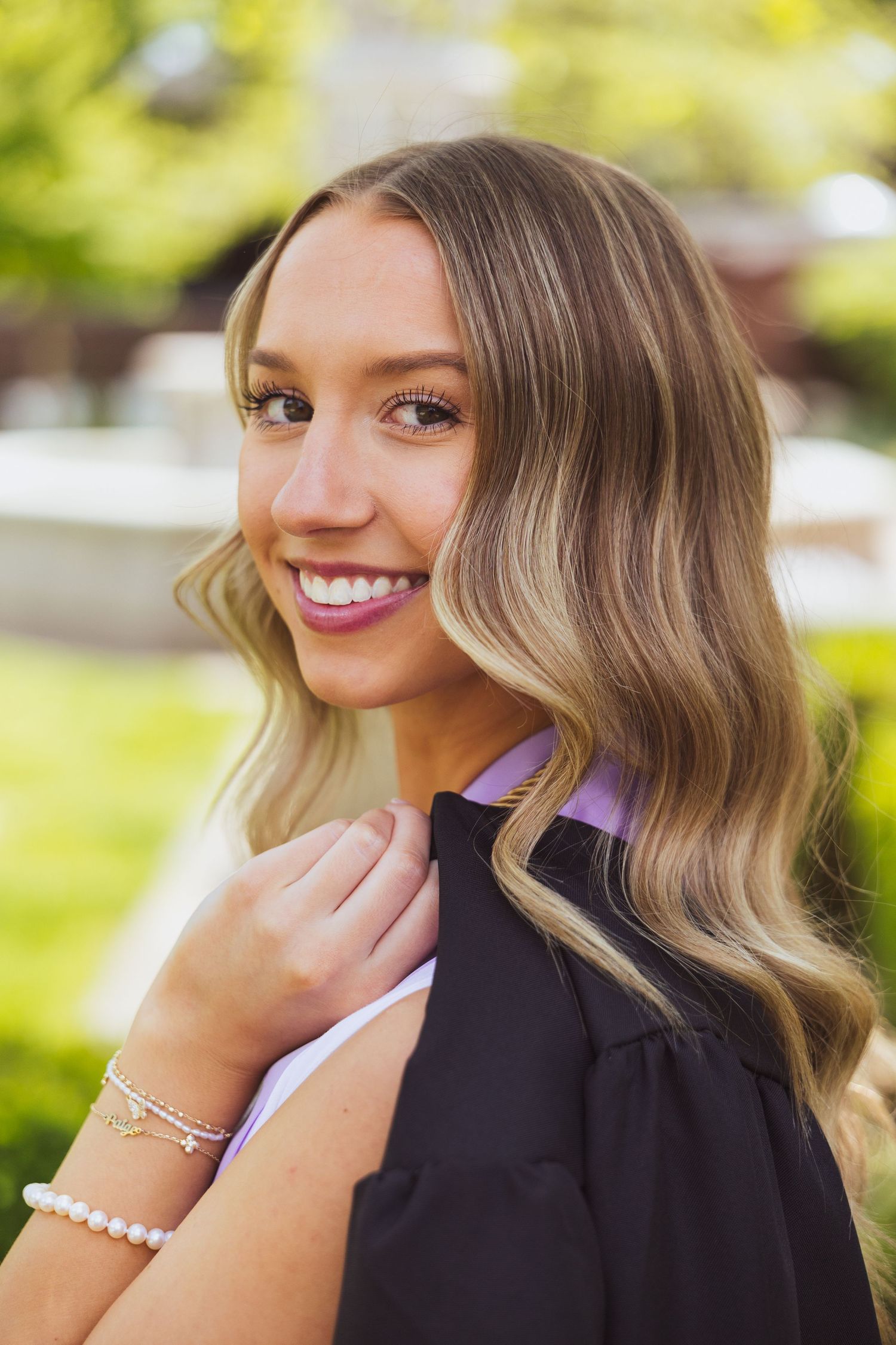 Portrait photo of someone with wavy blonde hair in a black top smiling outdoors with a blurred green background.