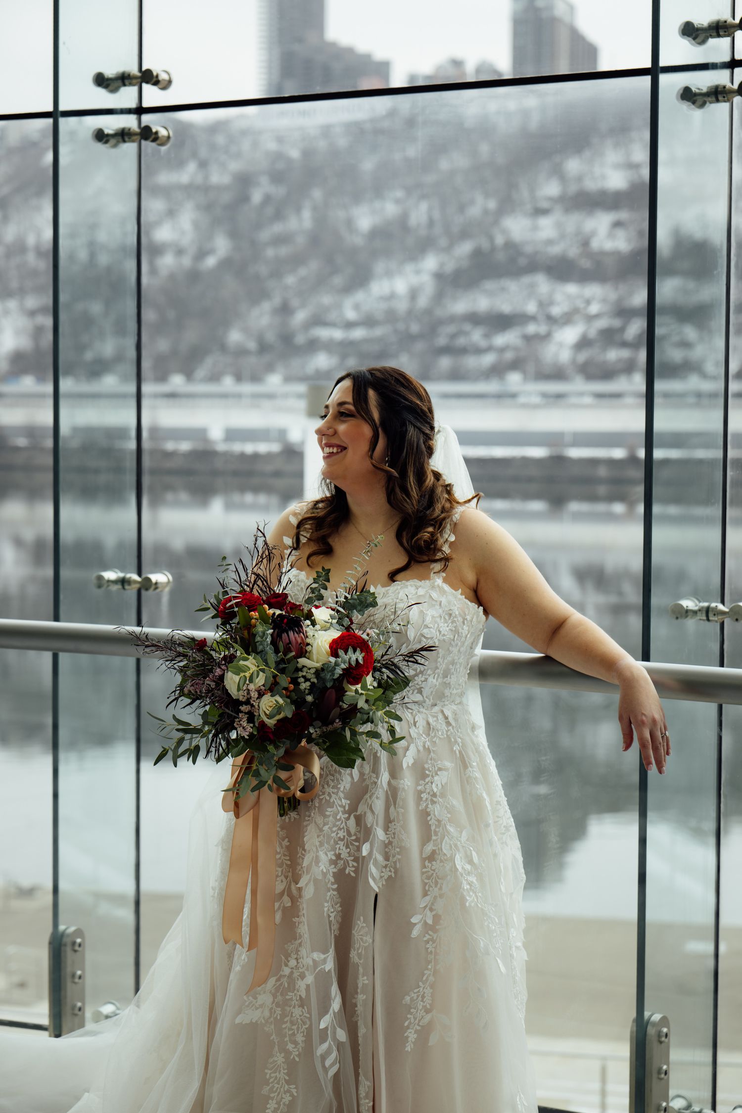 Smiling bride in lace wedding gown holds red and white bouquet by glass windows with snowy river view.