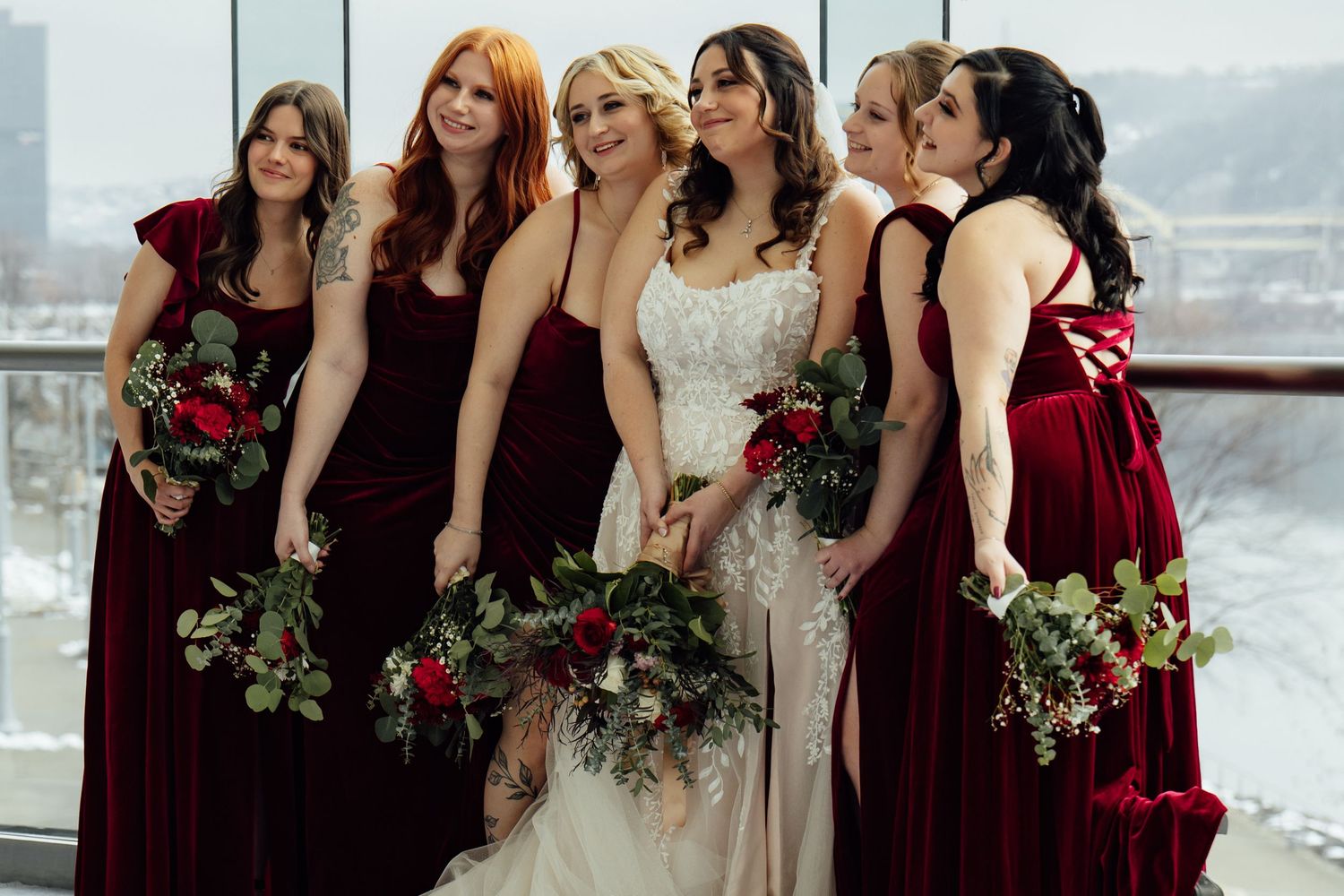 Bride in lace gown surrounded by five bridesmaids in burgundy velvet dresses holding red floral bouquets.