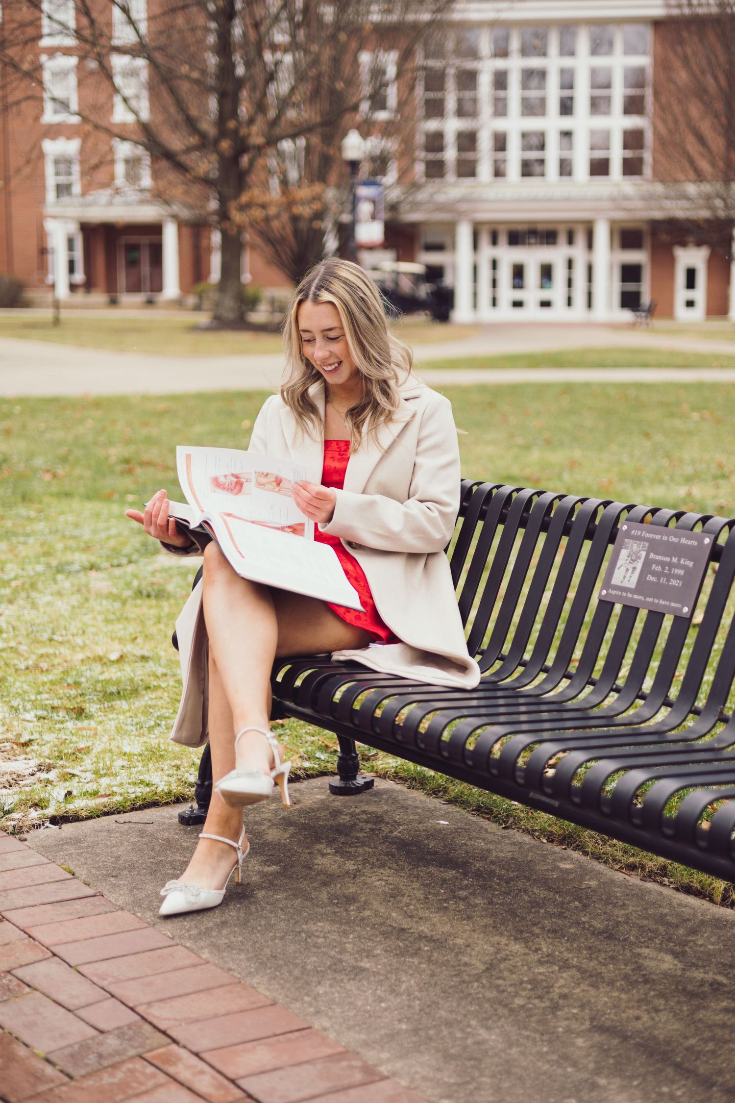 Graduate in red dress and cream coat reads while sitting on bench outside academic building.