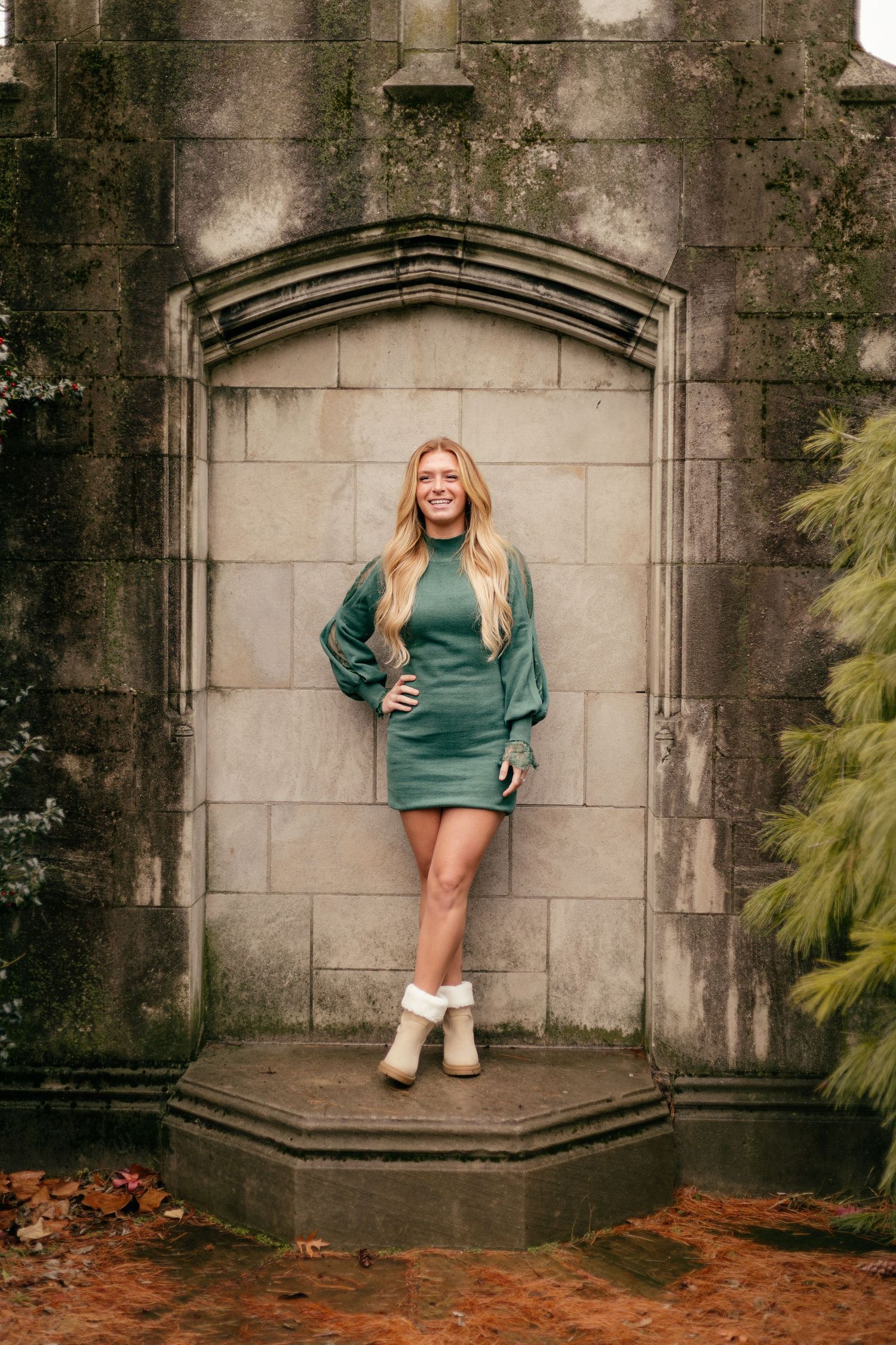Woman in green sweater dress and white boots poses confidently in gothic stone archway.