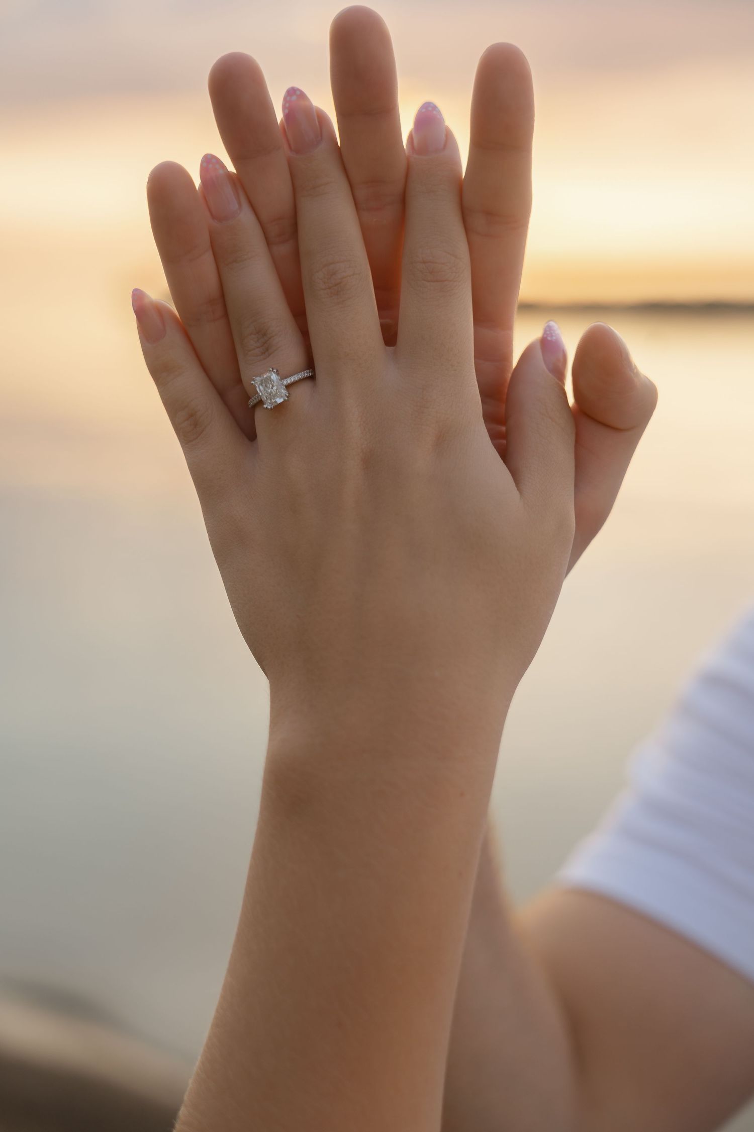 Two hands touching at sunset, one adorned with a sparkling diamond solitaire engagement ring on the ring finger.