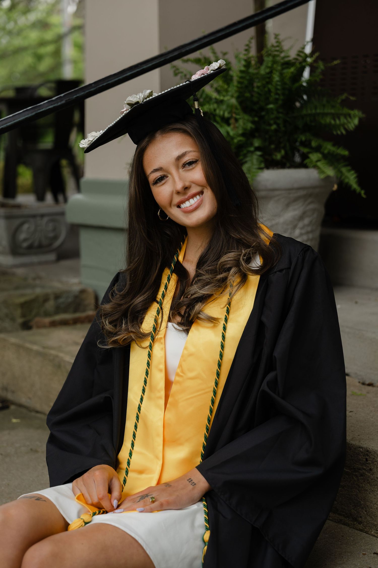 Smiling young woman in black graduation cap and gown with gold stole sitting on steps.