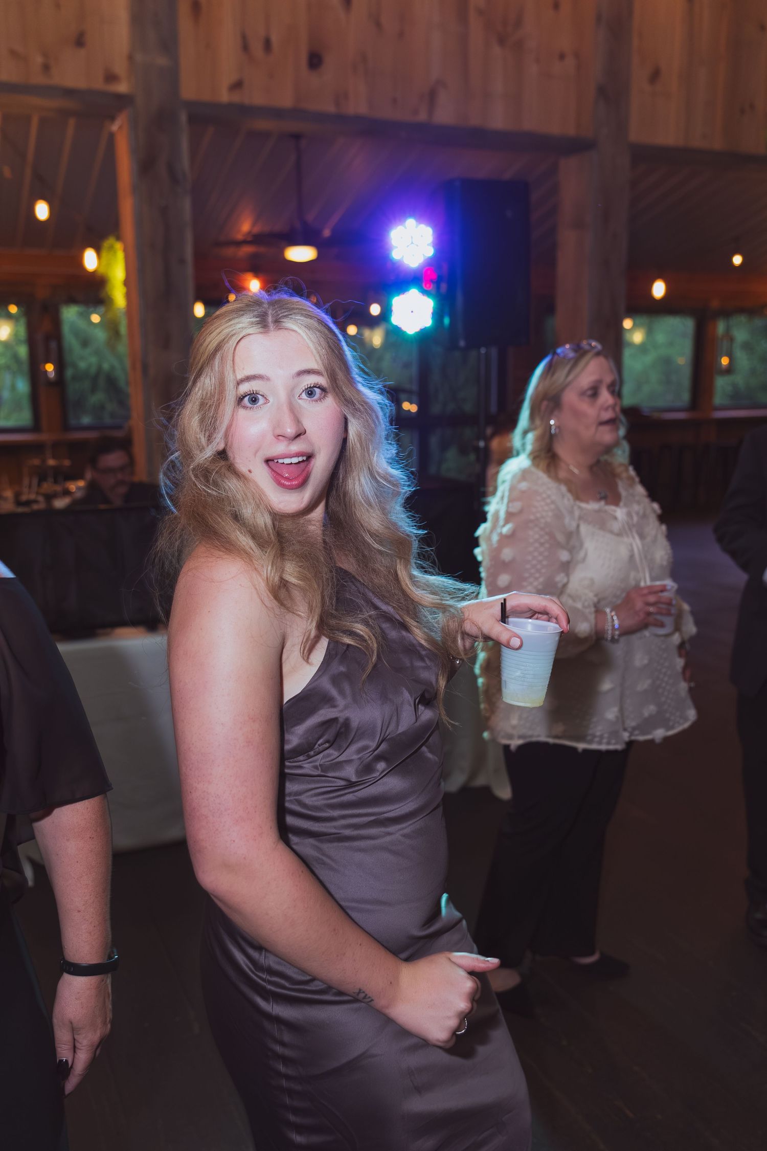 Blonde woman in gray satin dress smiling at wedding reception with colorful dance floor lights.