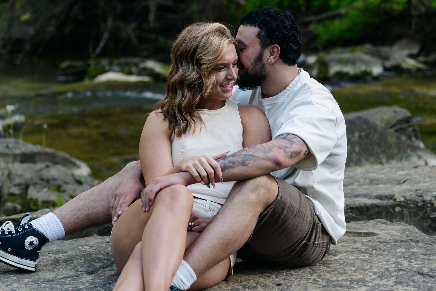 Couple sitting on rocks by a creek, sharing a romantic moment outdoors in nature.