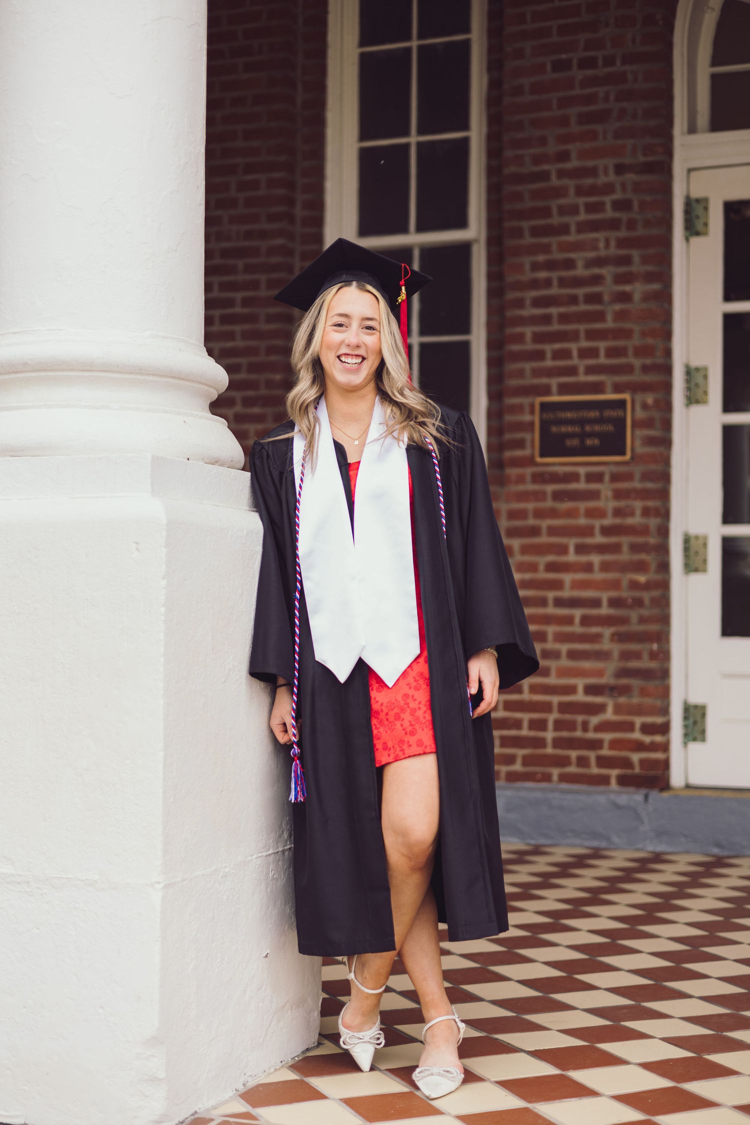 Graduate in cap and gown poses near brick building with checkered floor and white columns.