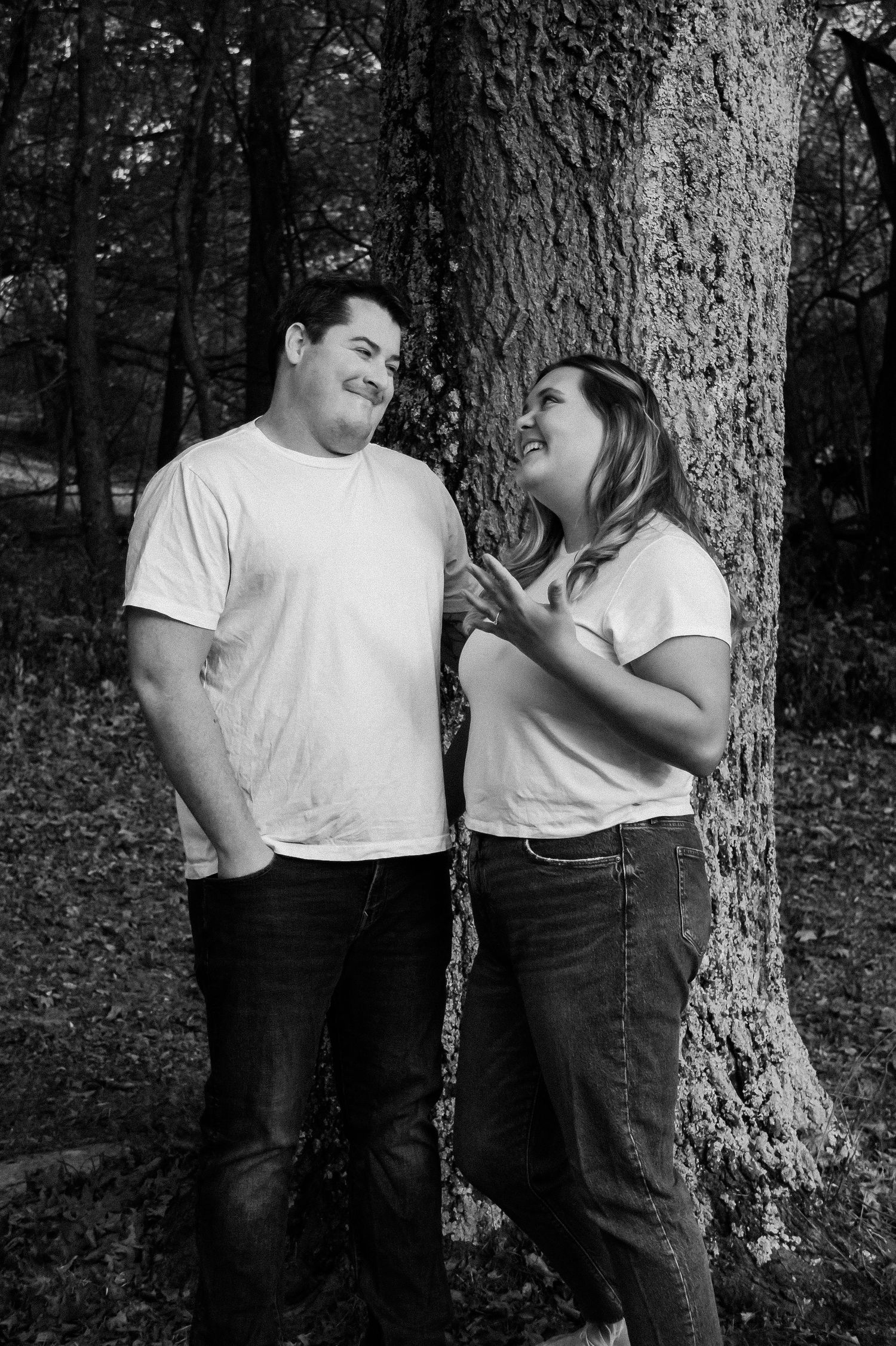 Black and white photo of couple sharing joyful moment by tree in woods.